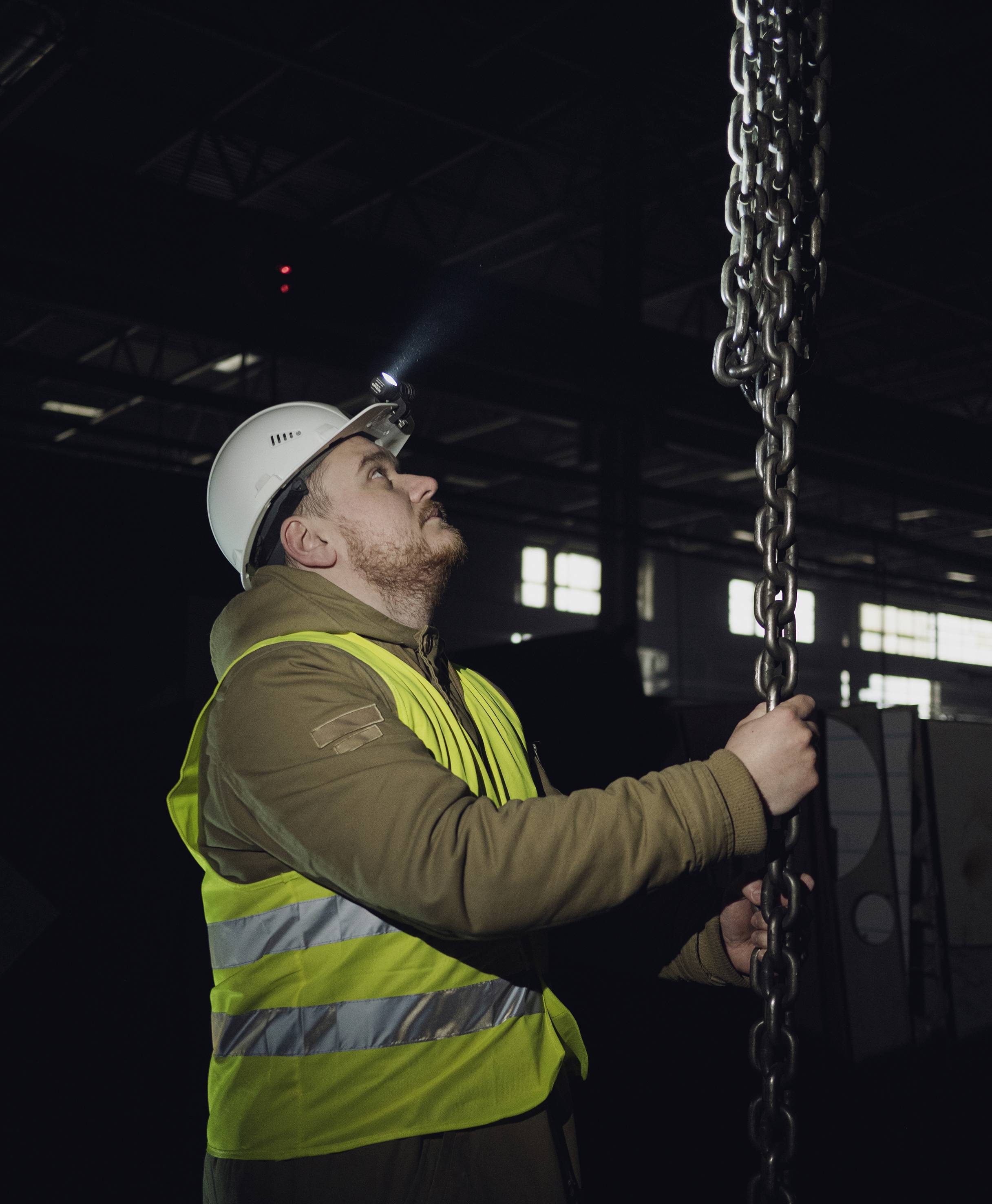 Ein Arbeiter mit Schutzhelm und Warnweste inspiziert eine schwere Kette in einer Fabrikhalle bei schwachem Licht.