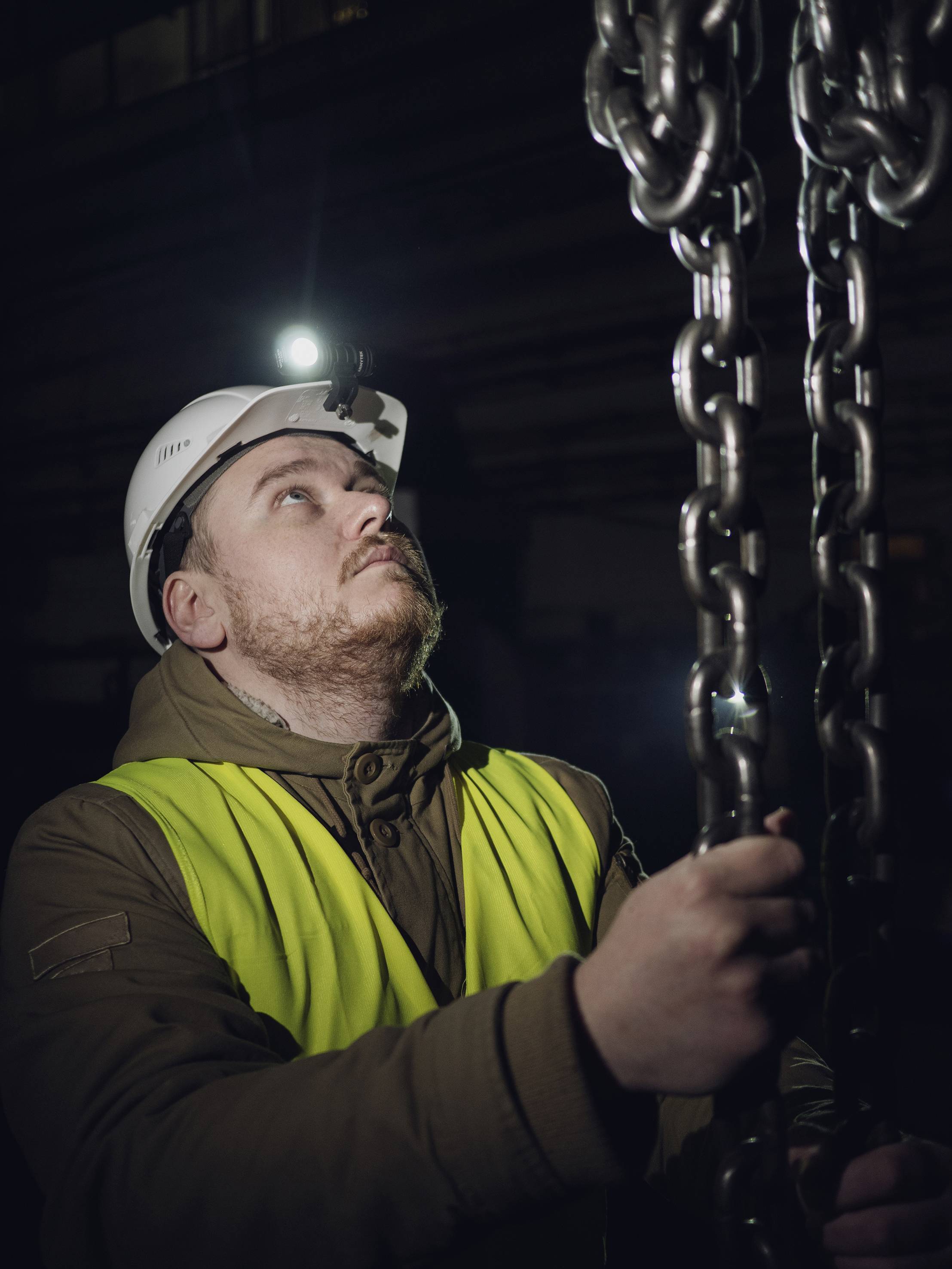 Ein Bauarbeiter in Sicherheitsweste mit Helm und Stirnlampe blickt auf eine schwere Kette. Dunkler Hintergrund deutet auf Nachtschicht hin.