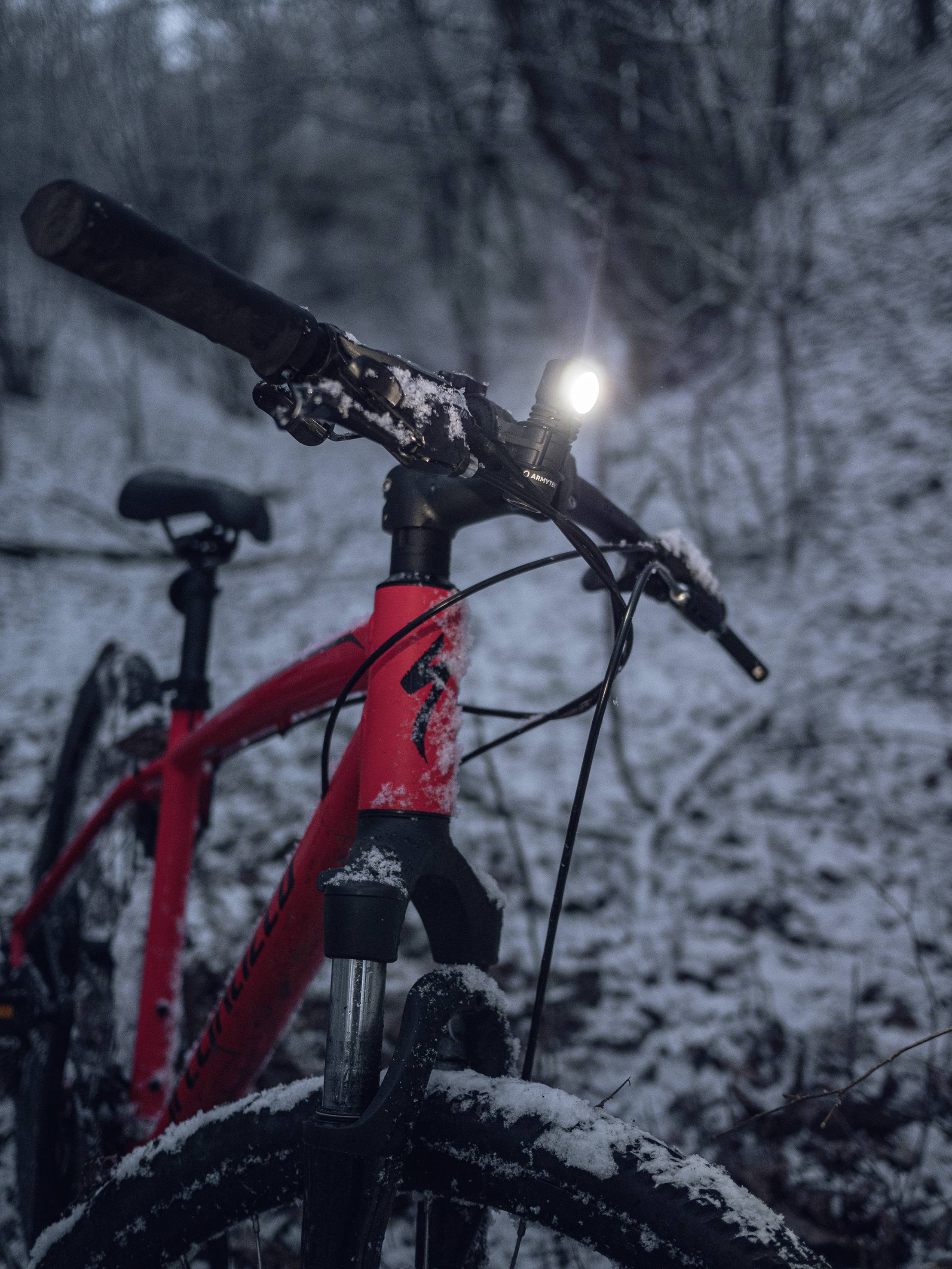 Rotes Fahrrad mit eingeschaltetem Vorderlicht steht im verschneiten Wald. Dunkle, winterliche Umgebung.