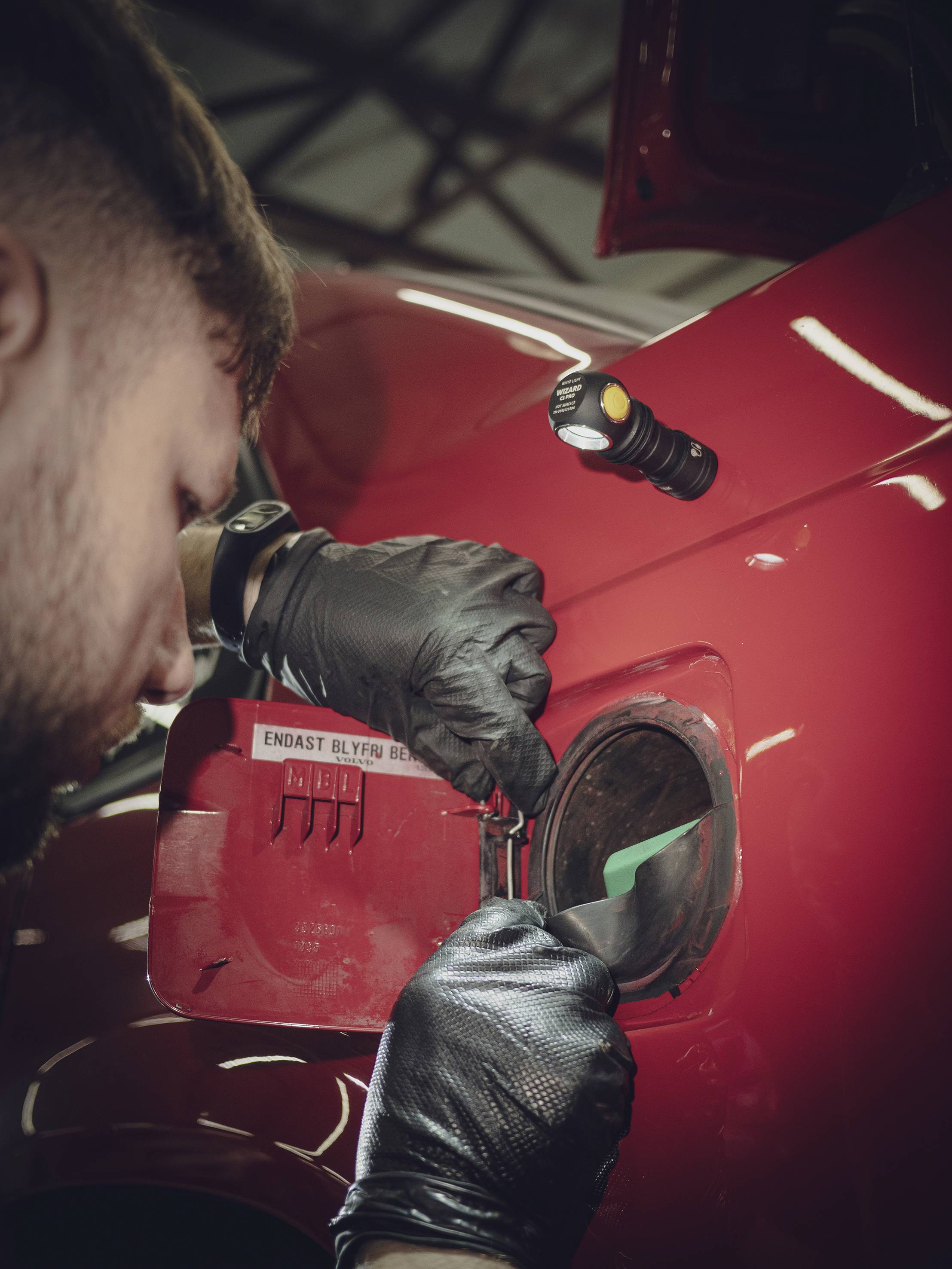 Mann mit schwarzen Handschuhen öffnet den Tankdeckel eines roten Fahrzeugs in einer Werkstattumgebung.