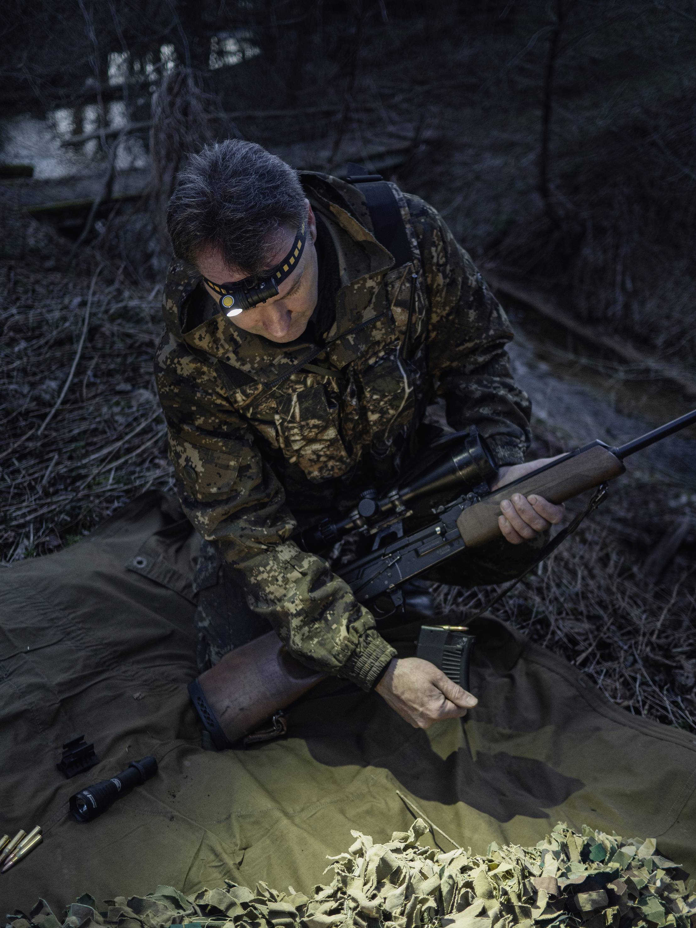 Eine Person in Tarnkleidung lädt ein Magazin in ein Gewehr in einem Waldgebiet bei Nacht, unterstützt von Stablampenlicht.