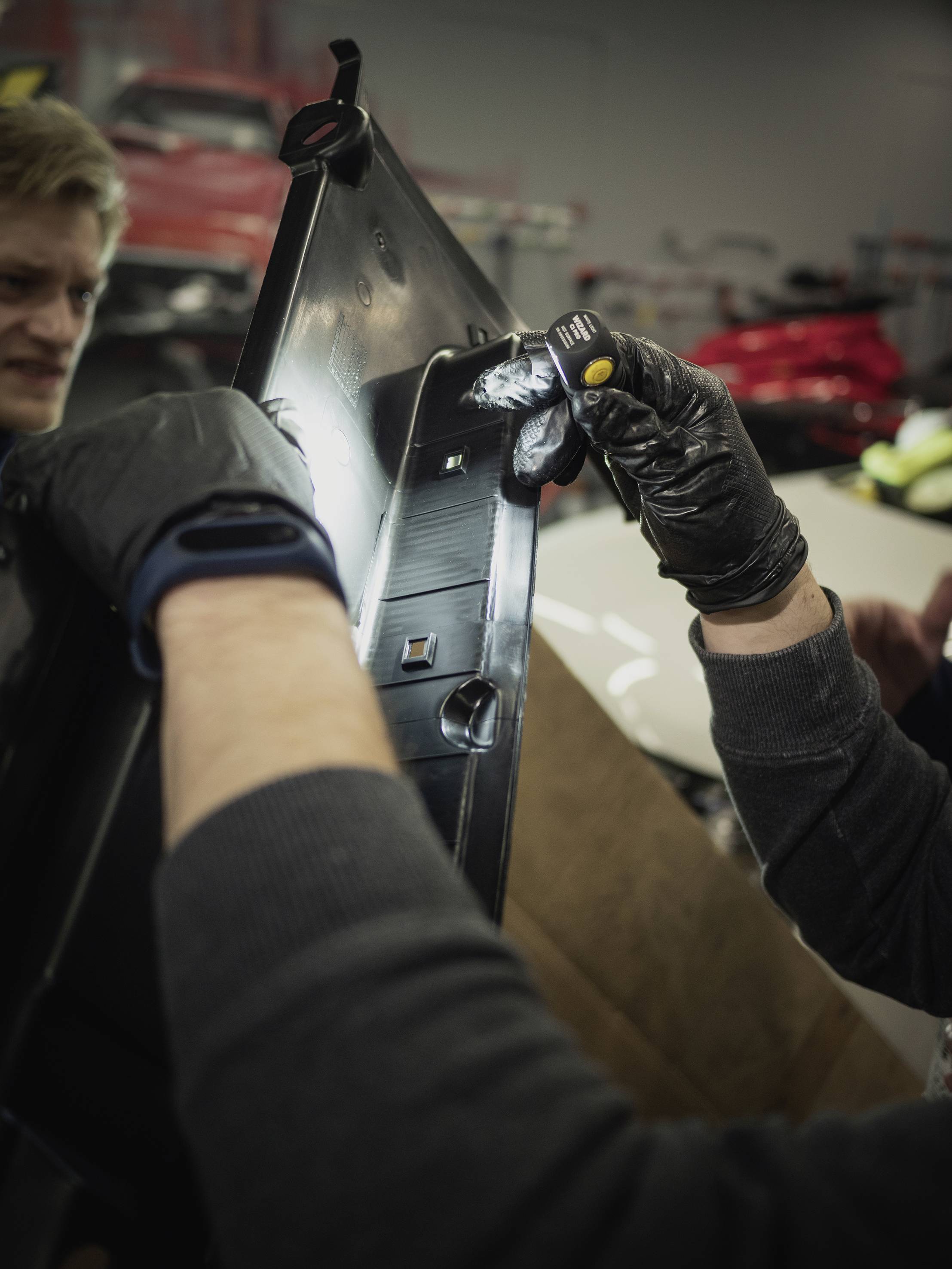 Zwei Personen arbeiten gemeinsam an einem Autoteil in einer Werkstatt, eine Person hält eine Taschenlampe und trägt Handschuhe.