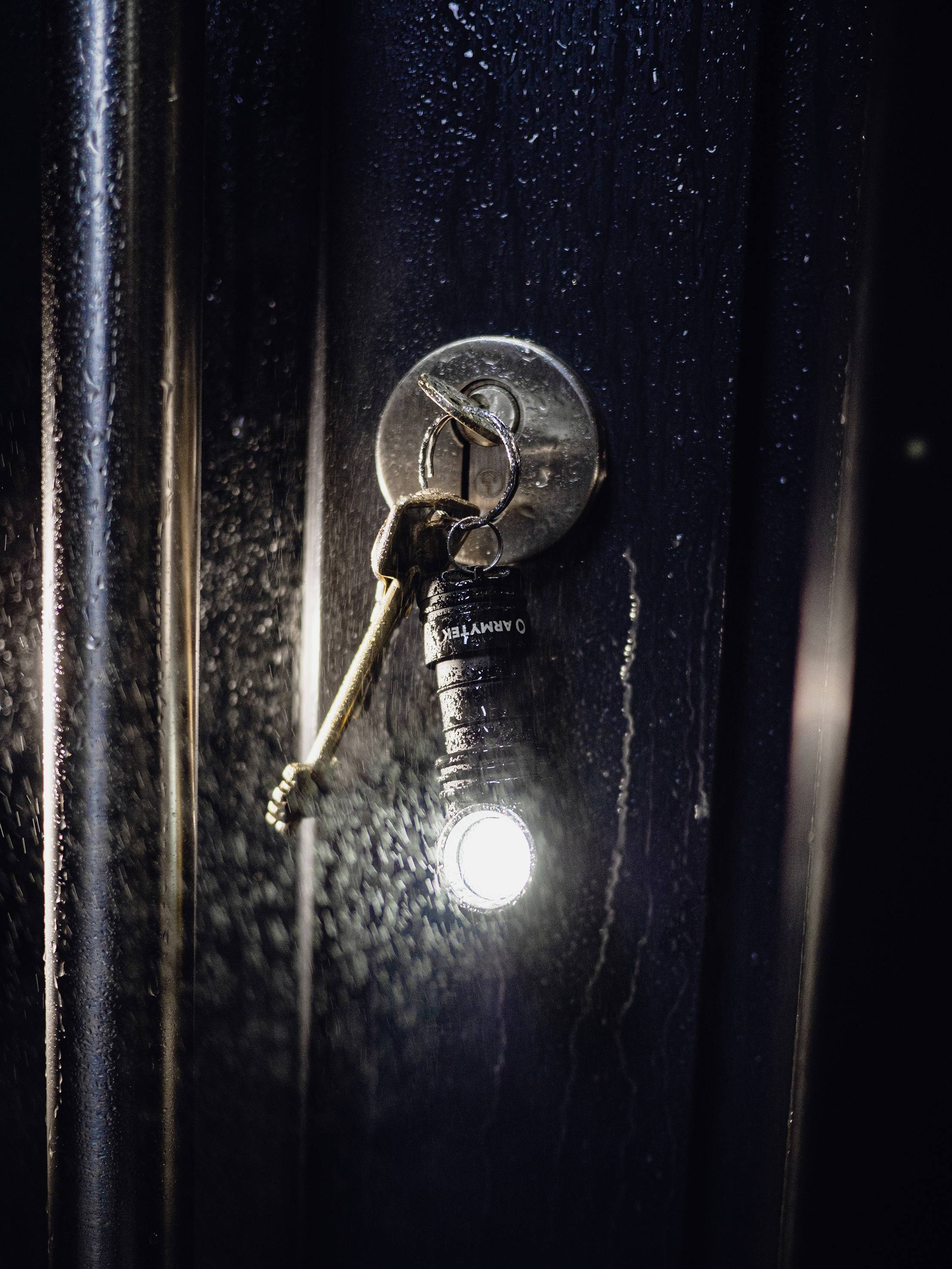 Schlüssel steckt in einem Türschloss, an dem eine leuchtende Mini-Taschenlampe hängt, beleuchtet die dunkle Tür.