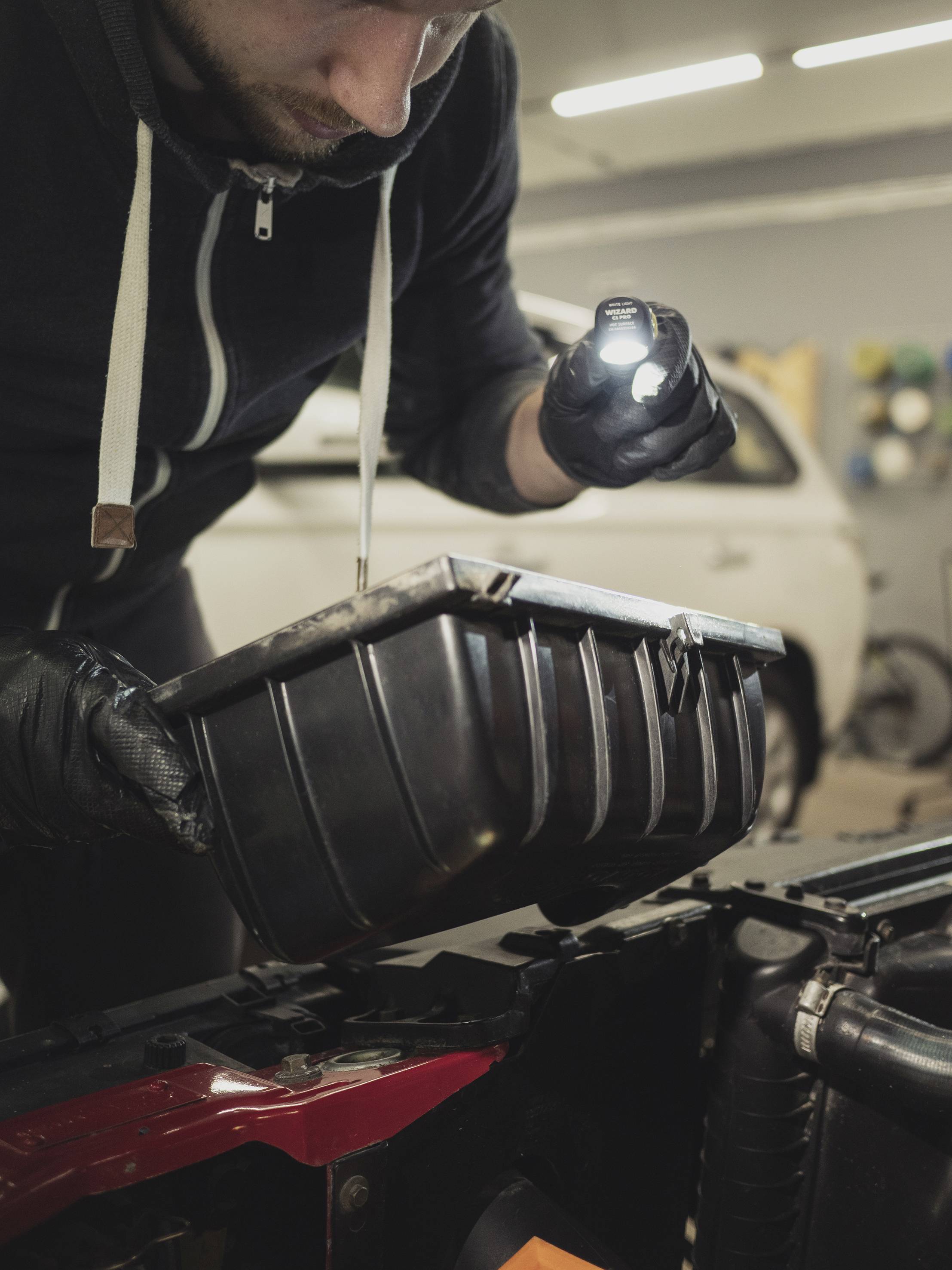 Eine Person überprüft ein Autoteil in einer Werkstatt, hält eine Taschenlampe und trägt schwarze Handschuhe. Im Hintergrund steht ein Auto.
