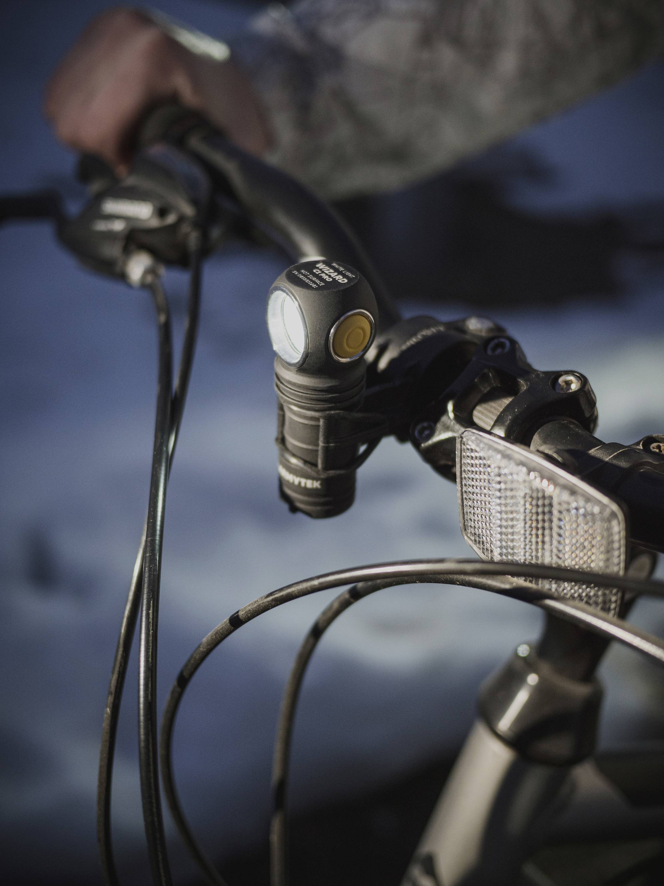 Eine Hand hält Fahrradlenker mit Frontlicht, umgeben von schneebedecktem Hintergrund. Vordergrund fokussiert auf Fahrrad und Licht.