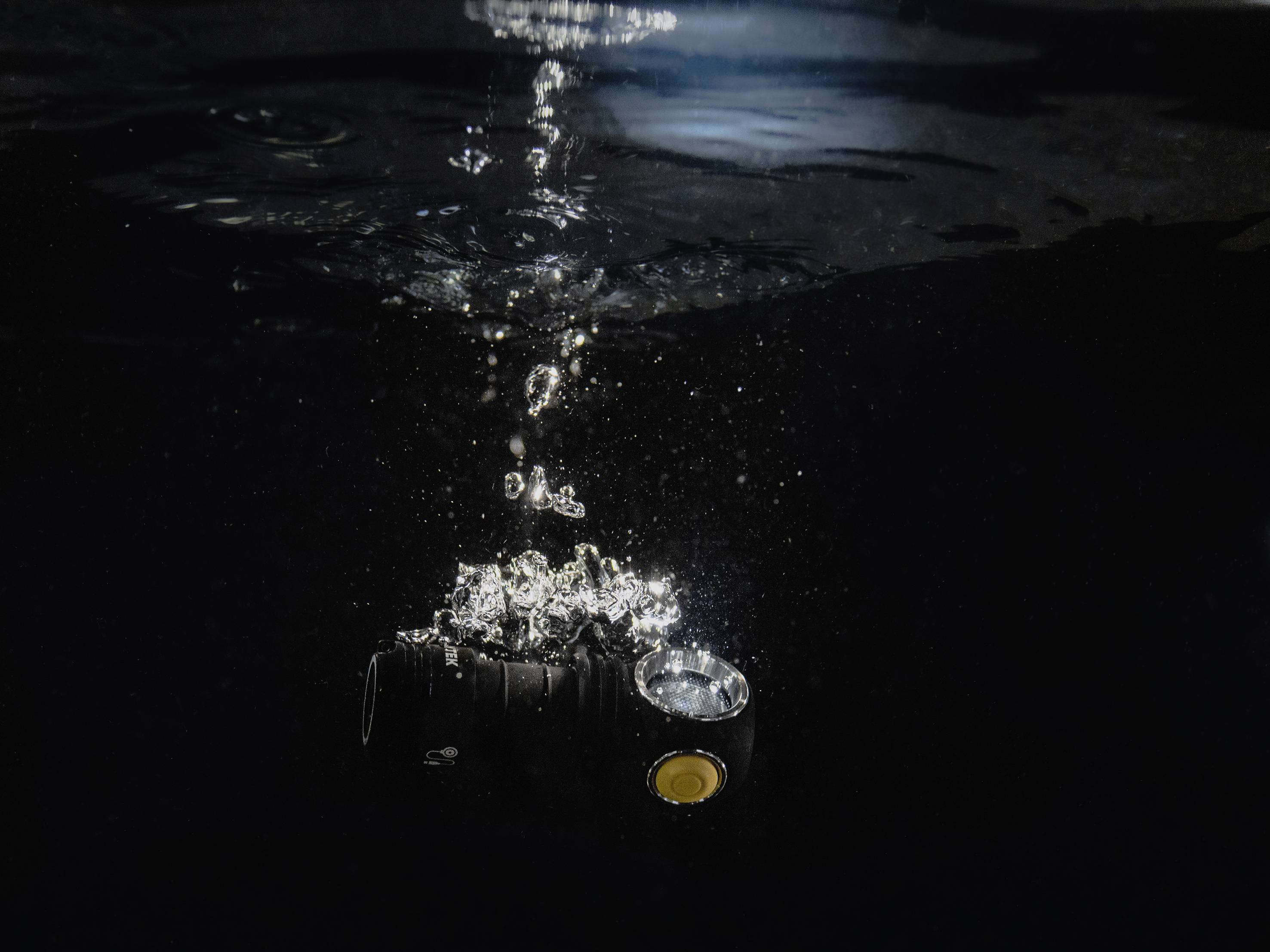 Eine wasserdichte, schwarze Taschenlampe leuchtet unter Wasser, während Blasen an die Oberfläche steigen. Die Szene ist dunkel beleuchtet.