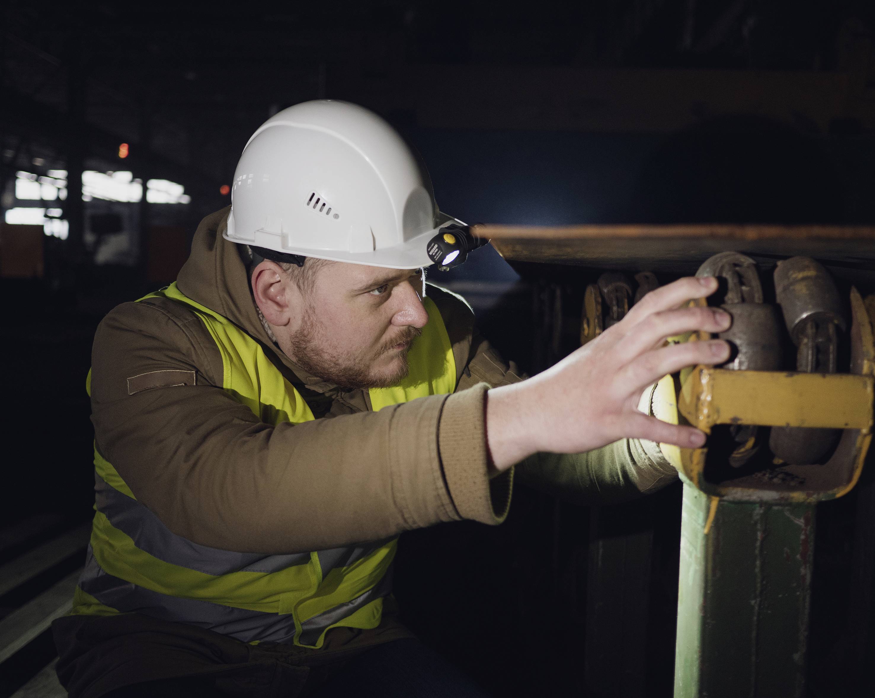 Ein Mann in Sicherheitskleidung und Helm inspiziert eine Maschine in einer dunklen industriellen Umgebung.