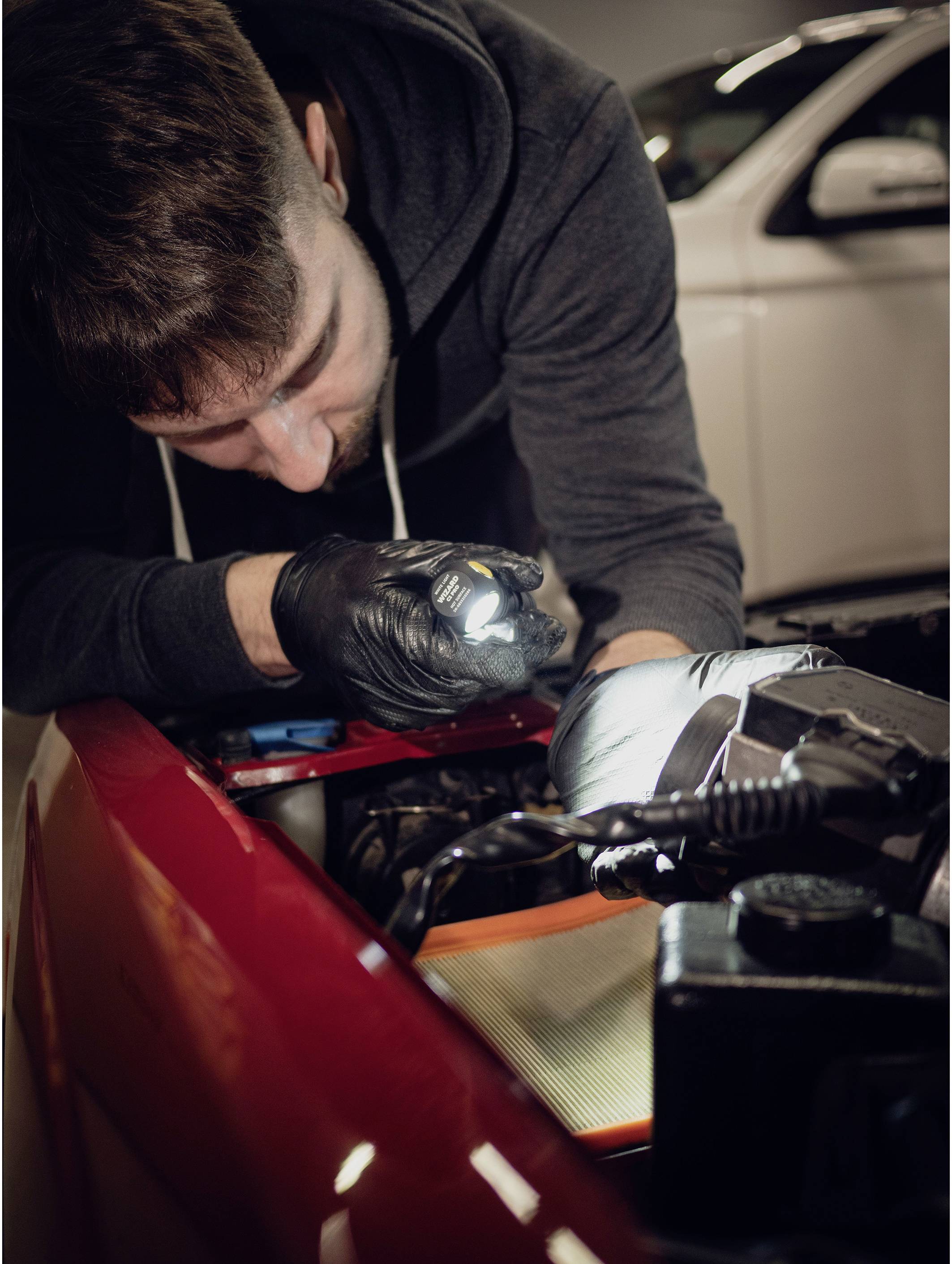 Mechaniker in schwarzem Hoodie und Handschuhen arbeitet mit Taschenlampe am Motor eines roten Autos in einer Werkstatt.