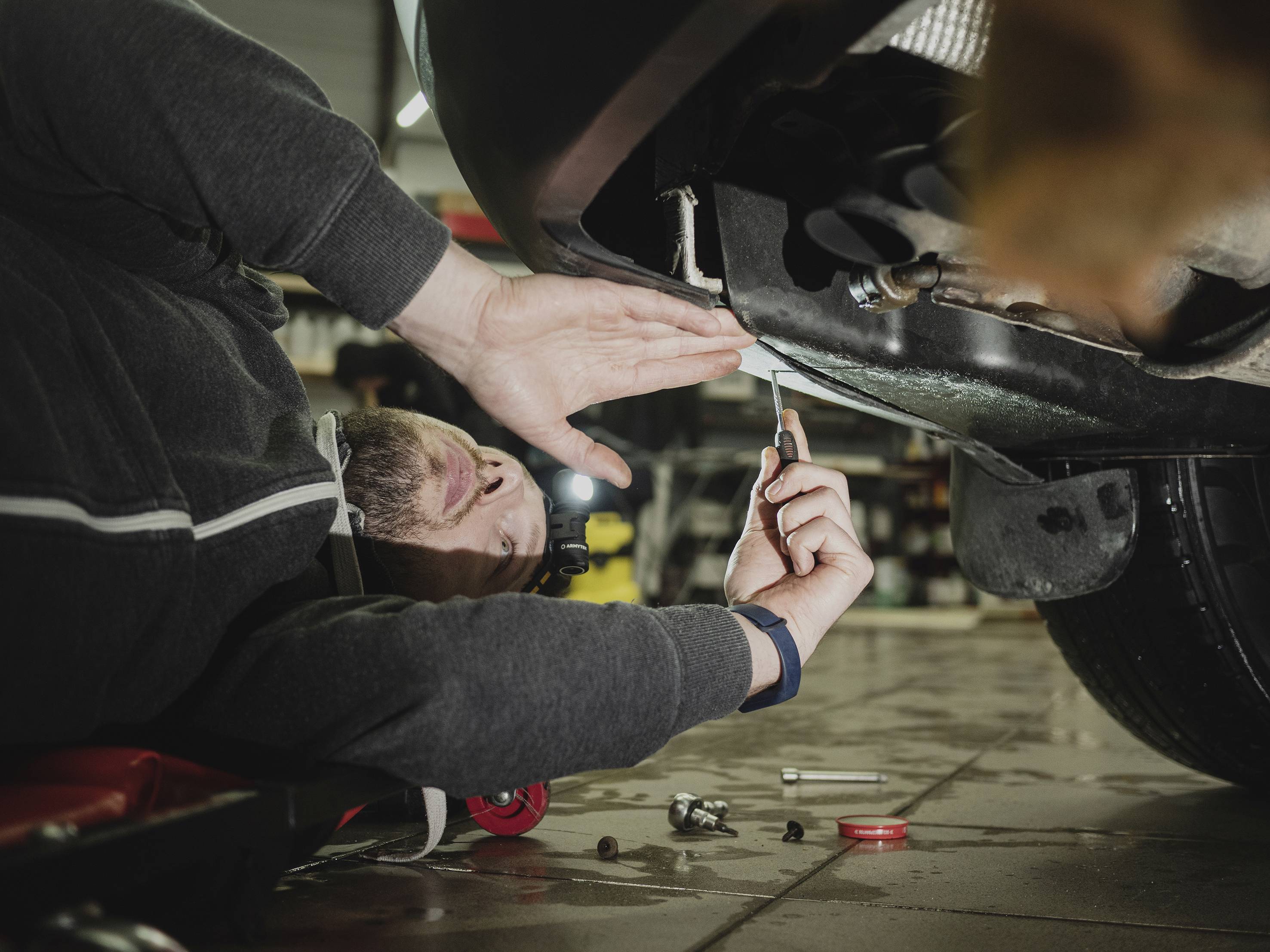 Ein Mechaniker liegt auf dem Rücken unter einem Auto und arbeitet mit einem Schraubenschlüssel an der Fahrzeugunterseite.