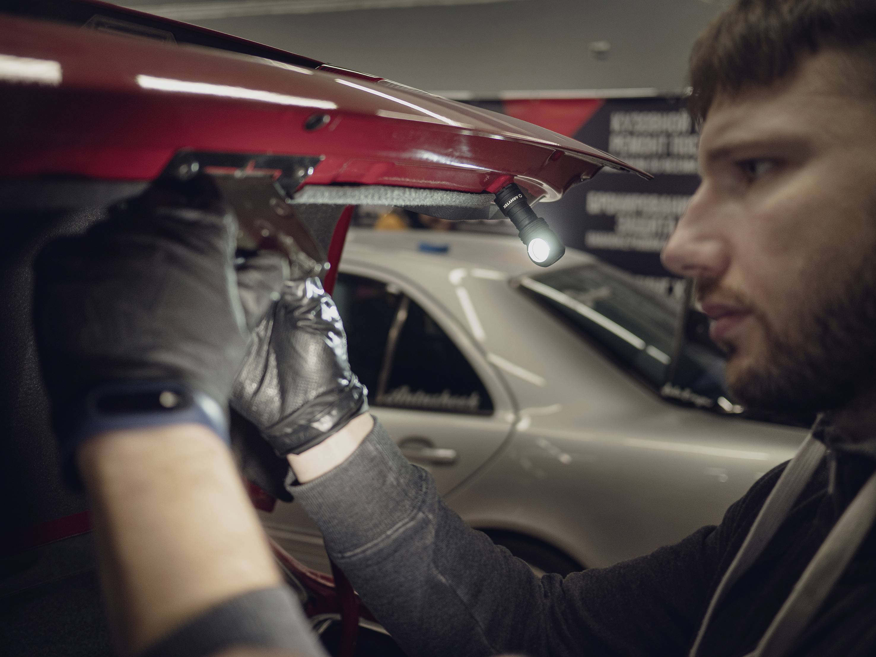 Ein Mann mit Handschuhen arbeitet konzentriert an einem Autoteil. Im Hintergrund ist unscharf ein Auto zu sehen.