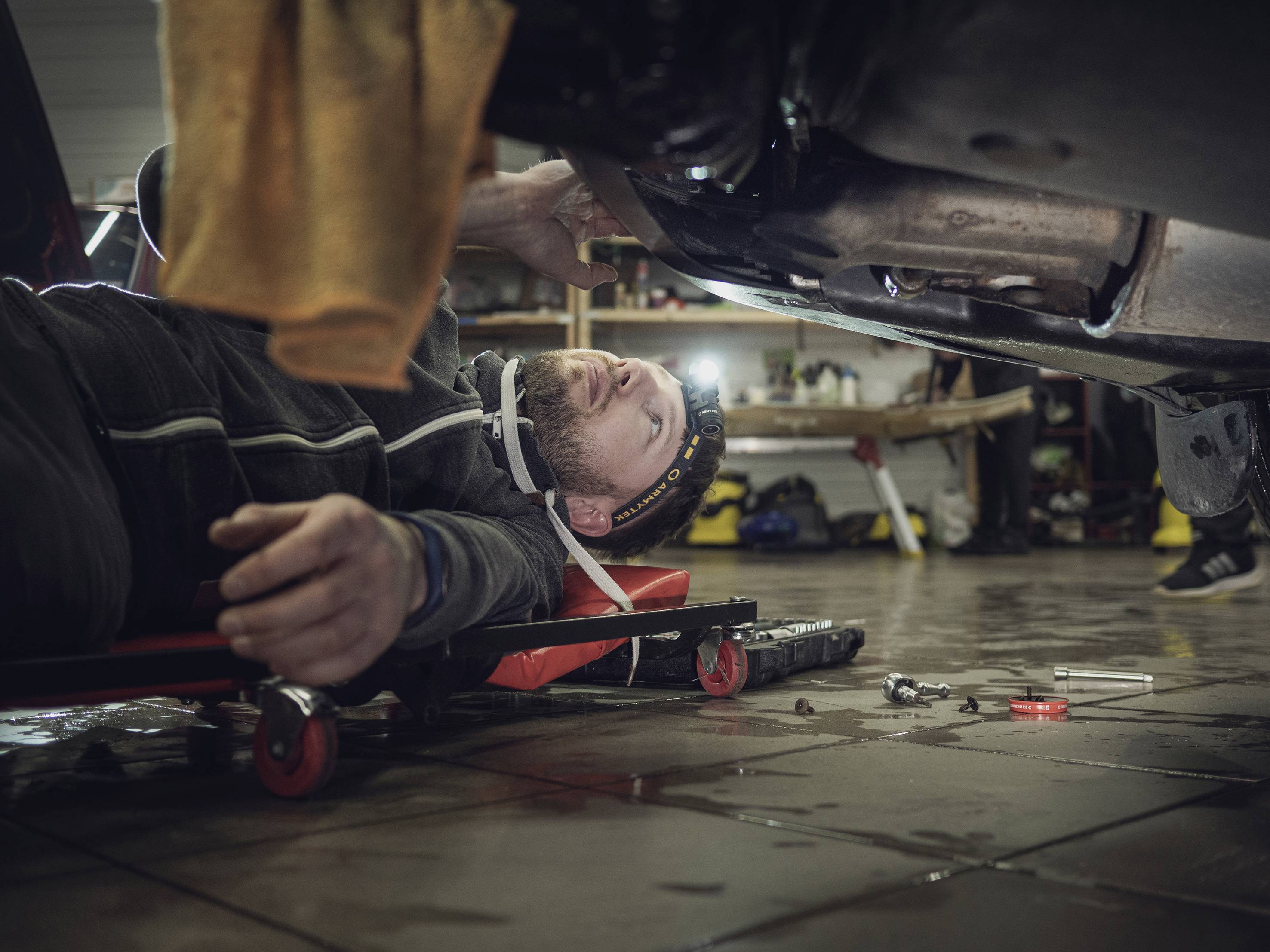 Ein Mechaniker liegt auf einem Rollbrett unter einem Auto in einer Werkstatt und arbeitet mit Werkzeugen an der Unterseite des Fahrzeugs.