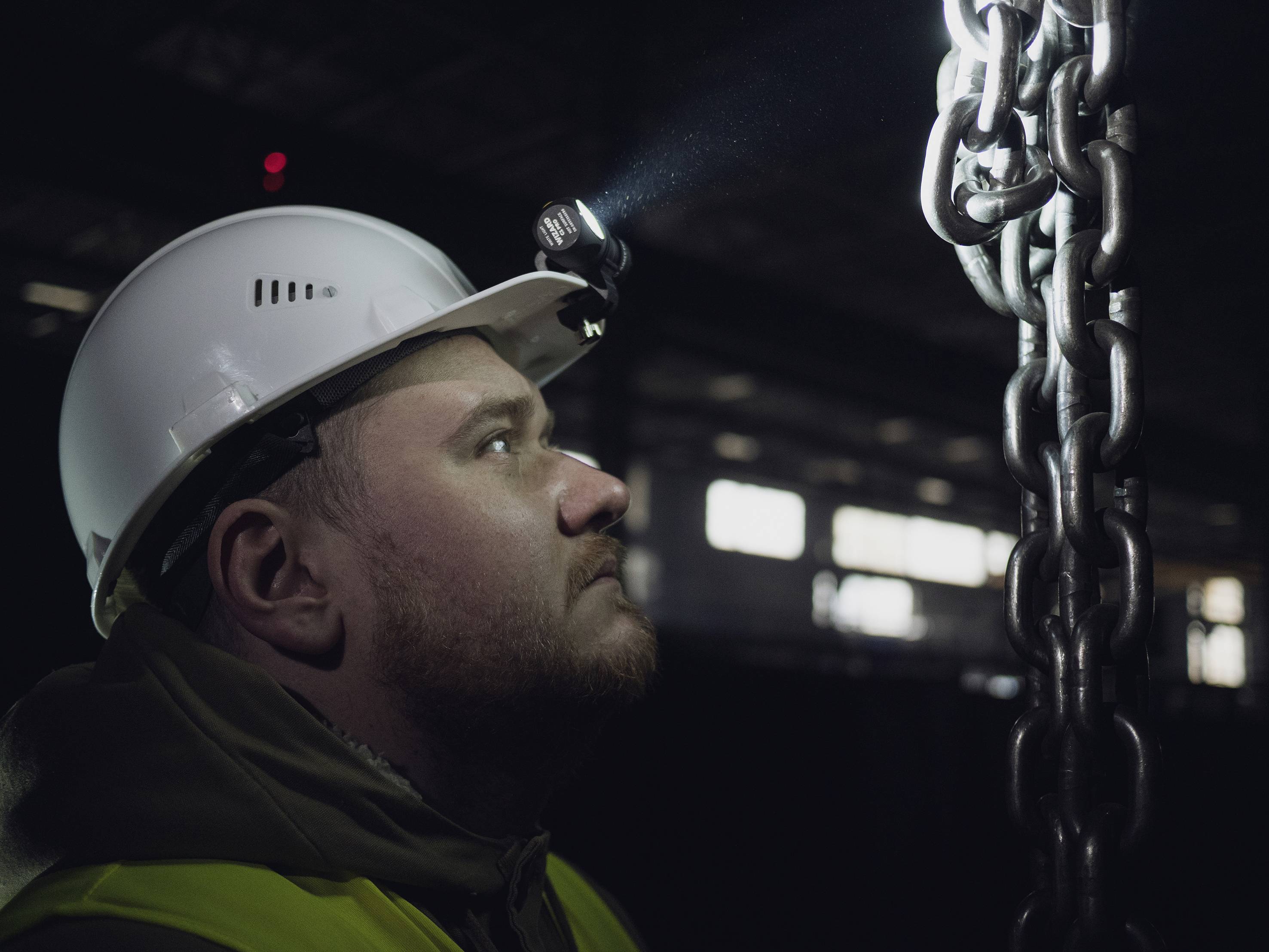 Ein Arbeiter mit Helm und Stirnlampe inspiziert sorgfältig eine schwere Metallkette in einem dunklen Industriegebäude.