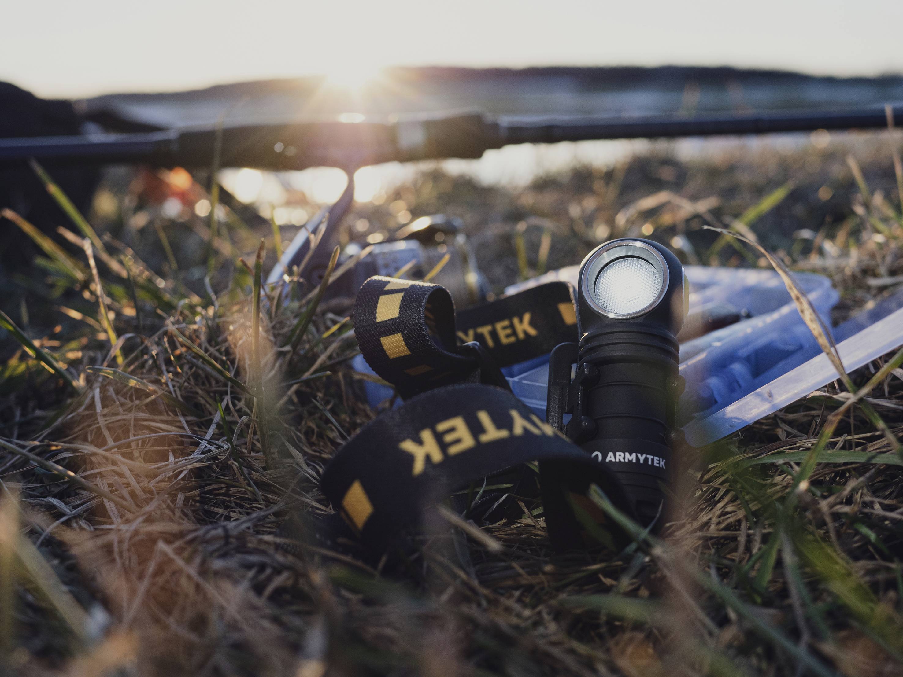Taschenlampe und Zubehör liegen auf grasigem Boden bei Sonnenuntergang, nahe eines Gewässers. Stimmungsvolle Outdoor-Szene.