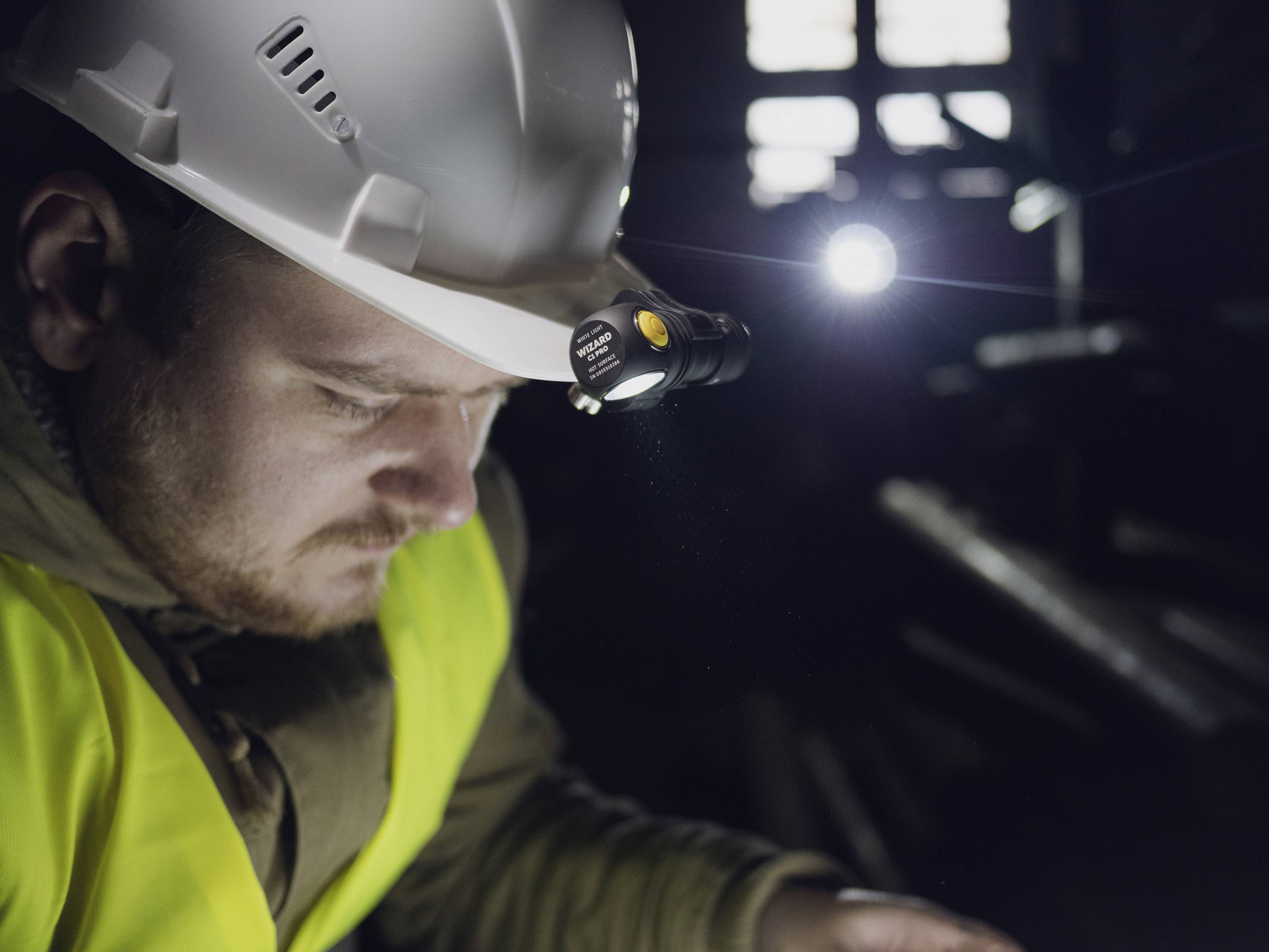 Ein Bauarbeiter mit Schutzhelm und Warnweste arbeitet in einem schwach beleuchteten Raum. Eine Stirnlampe leuchtet den Bereich aus.