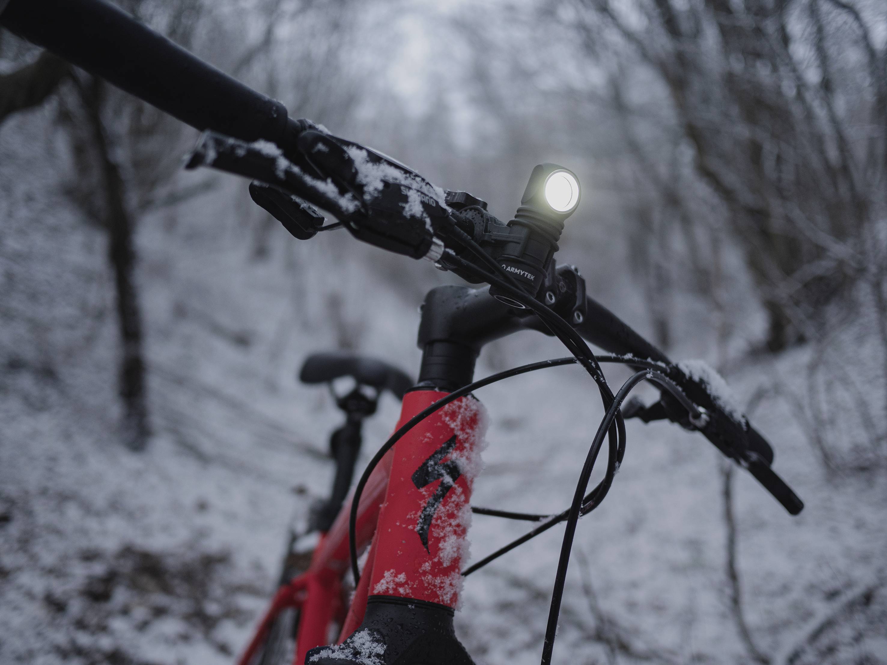 Ein rotes Fahrrad mit eingeschaltetem Licht steht in einem verschneiten Wald. Die Umgebung ist winterlich und bewaldet.