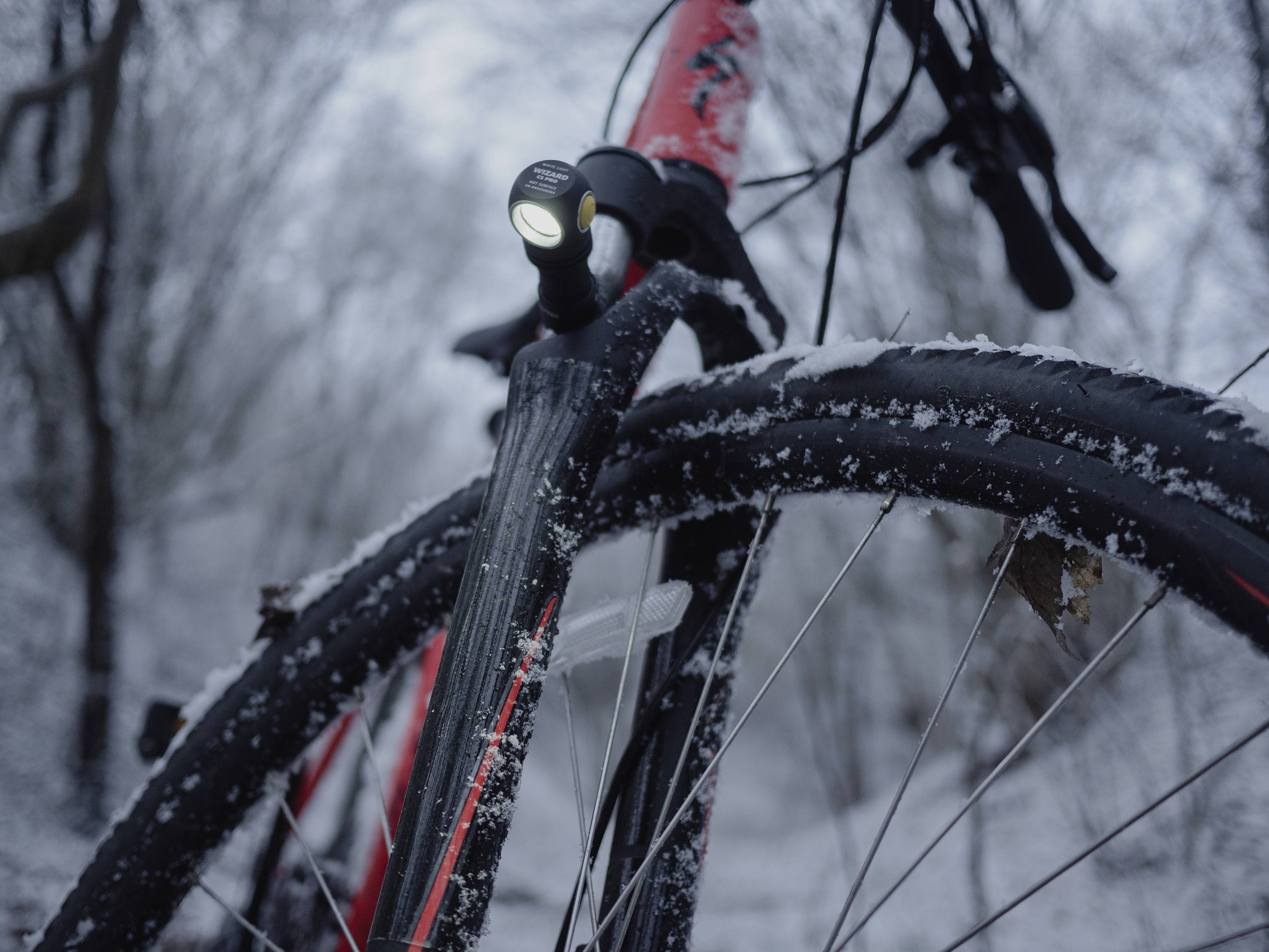 Fahrrad im Schnee mit eingeschaltetem Vorderlicht, umgeben von winterlicher Landschaft. Hauptfokus auf Vorderreifen und Lampe.