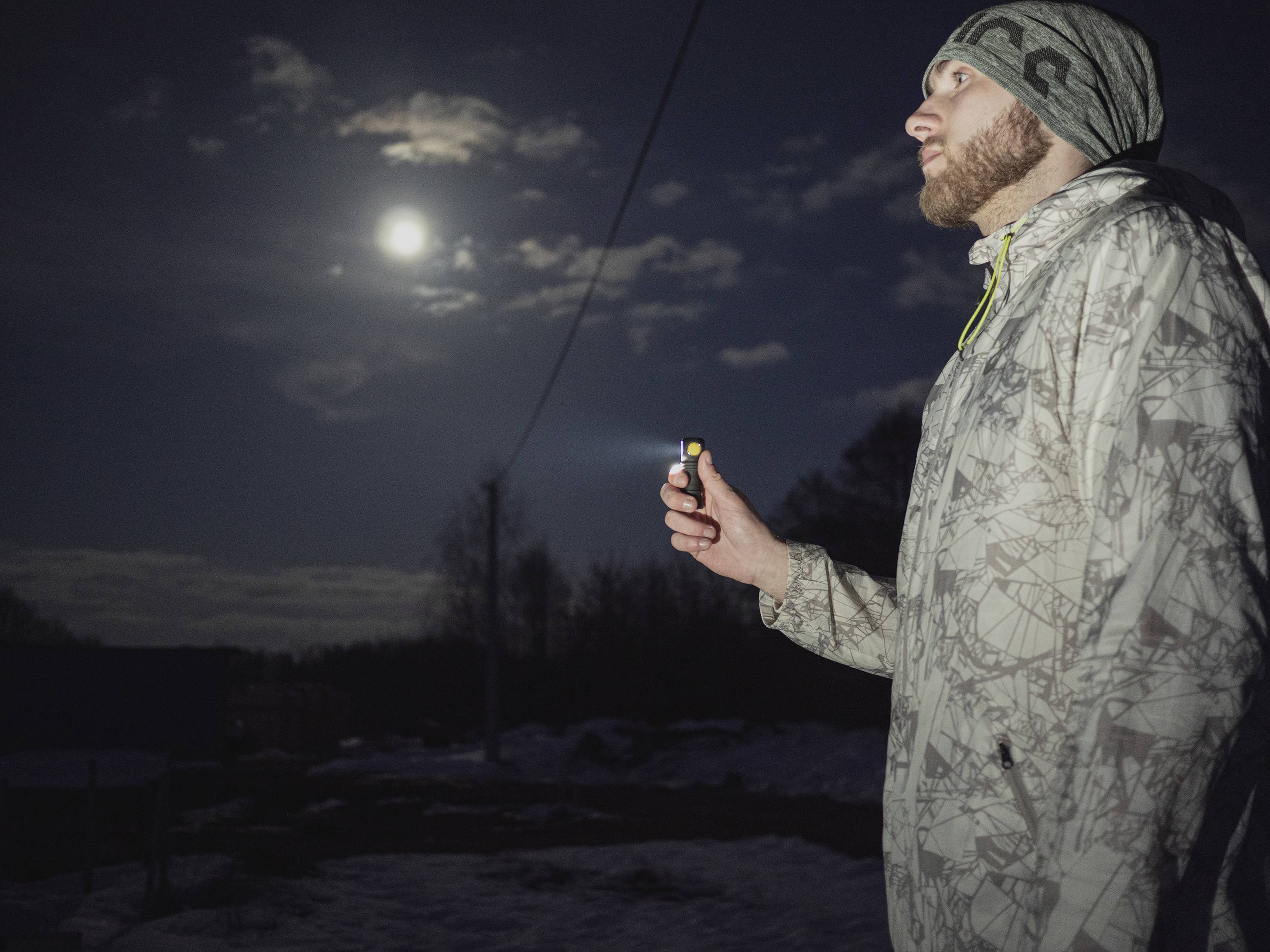 Eine Person im Wintermantel hält eine leuchtende Taschenlampe in der Nacht, während der Vollmond am bewölkten Himmel scheint.