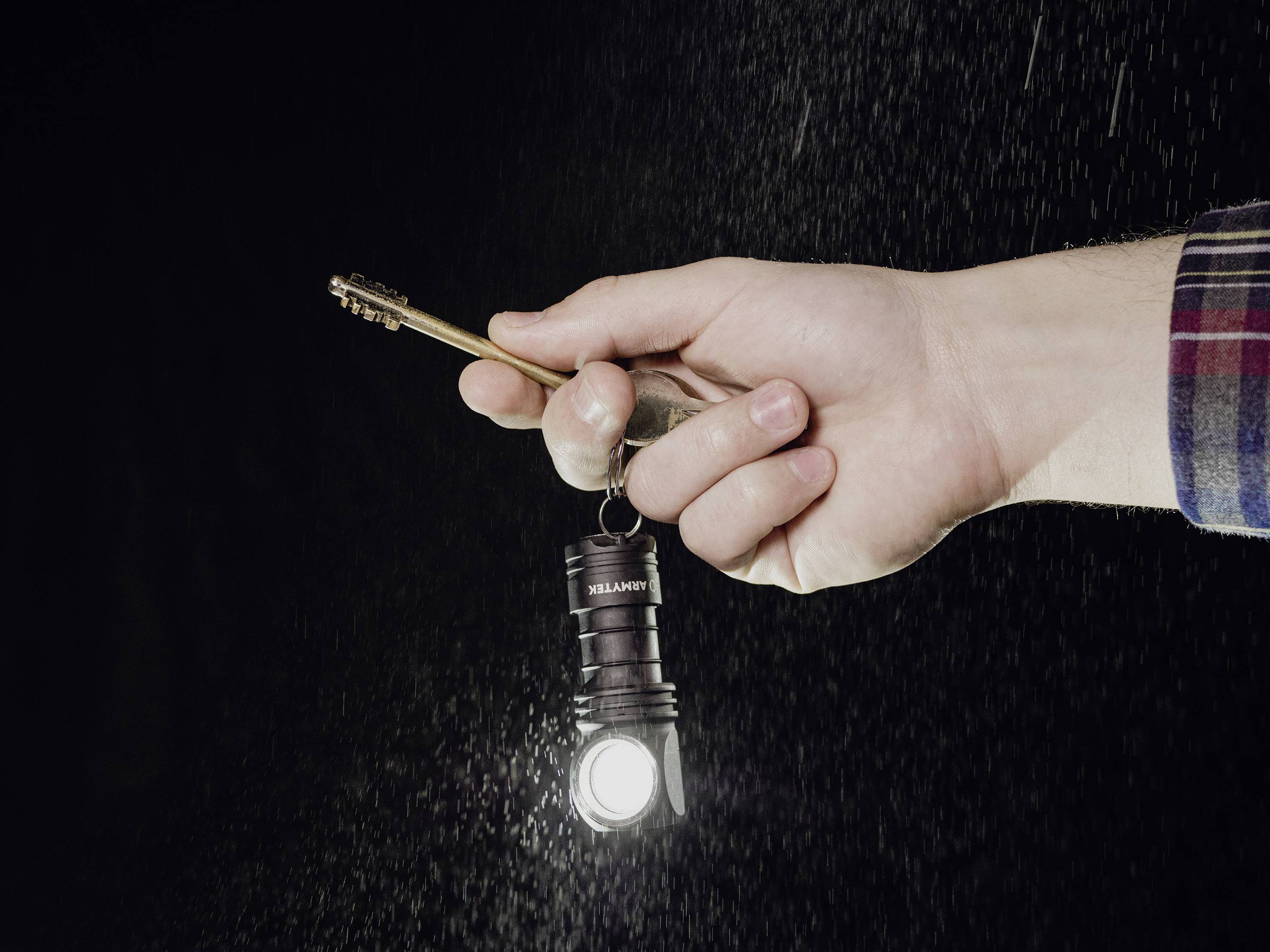 Eine Hand hält einen Schlüssel, an dem eine kleine Taschenlampe befestigt ist, die leuchtet. Hintergrund dunkel, möglicherweise regnet es.