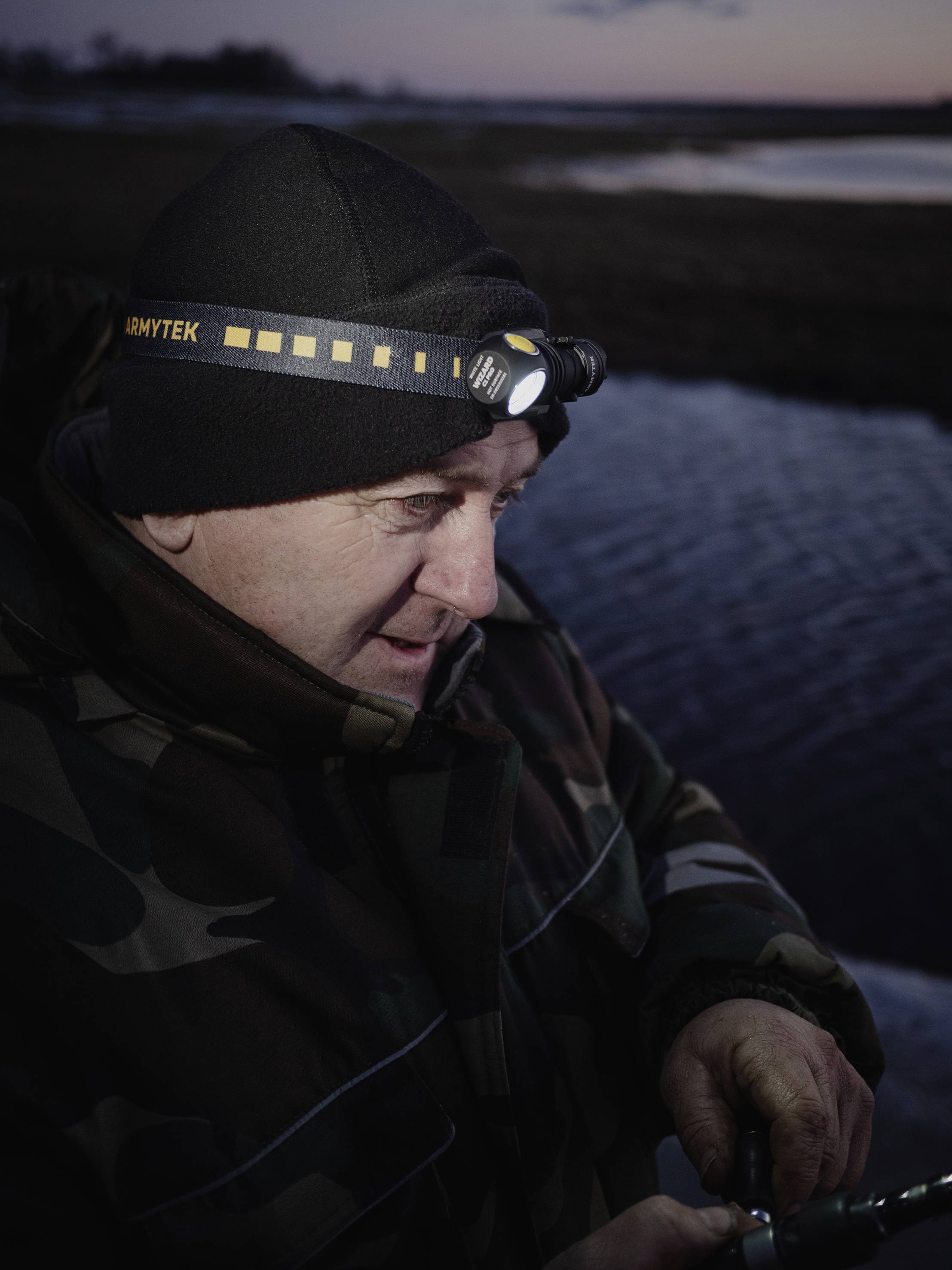 Person mit Stirnlampe angelt im Freien bei Dämmerung, warm eingepackt in Camouflage-Jacke. Hintergrund zeigt Wasser und dunklen Himmel.