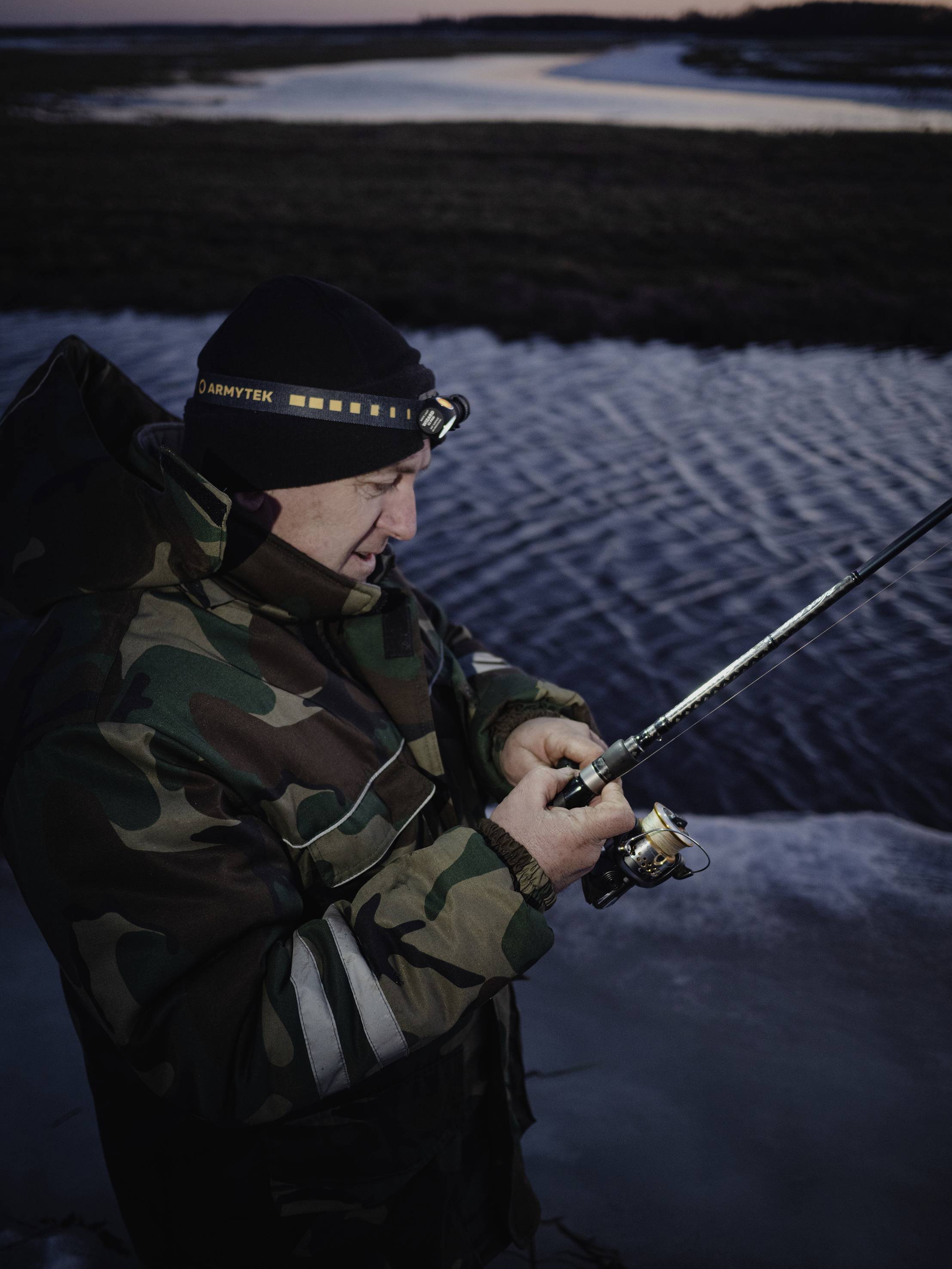 Eine Person in Tarnkleidung angelt bei Dämmerung an einem Fluss. Sie trägt eine Stirnlampe und konzentriert sich auf die Angelrute.