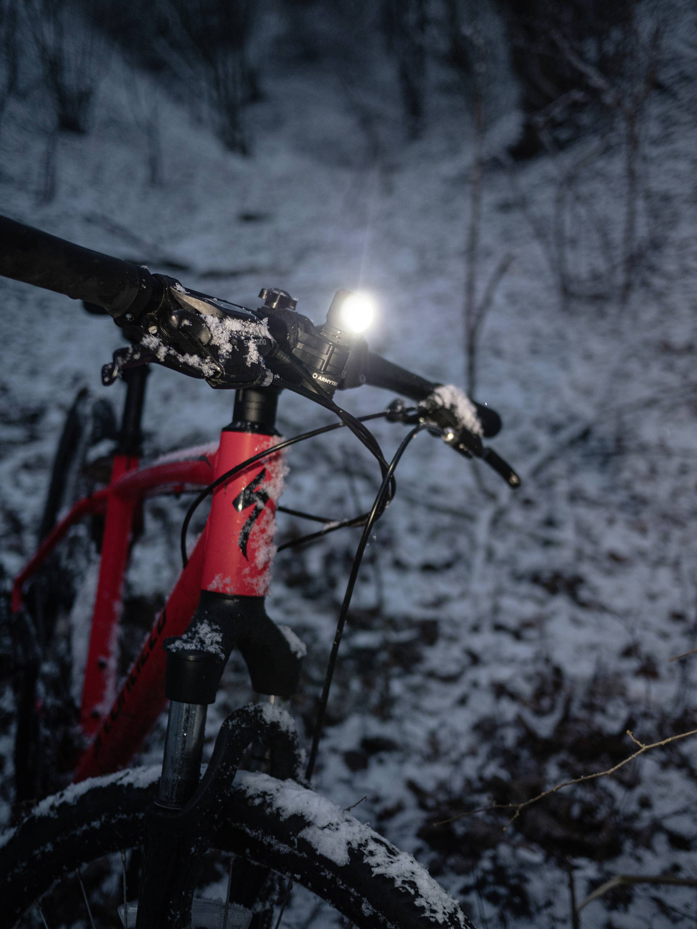 Rotes Fahrrad mit eingeschaltetem Frontlicht steht im schneebedeckten Wald. Die Umgebung ist dunkel, nur das Licht leuchtet.