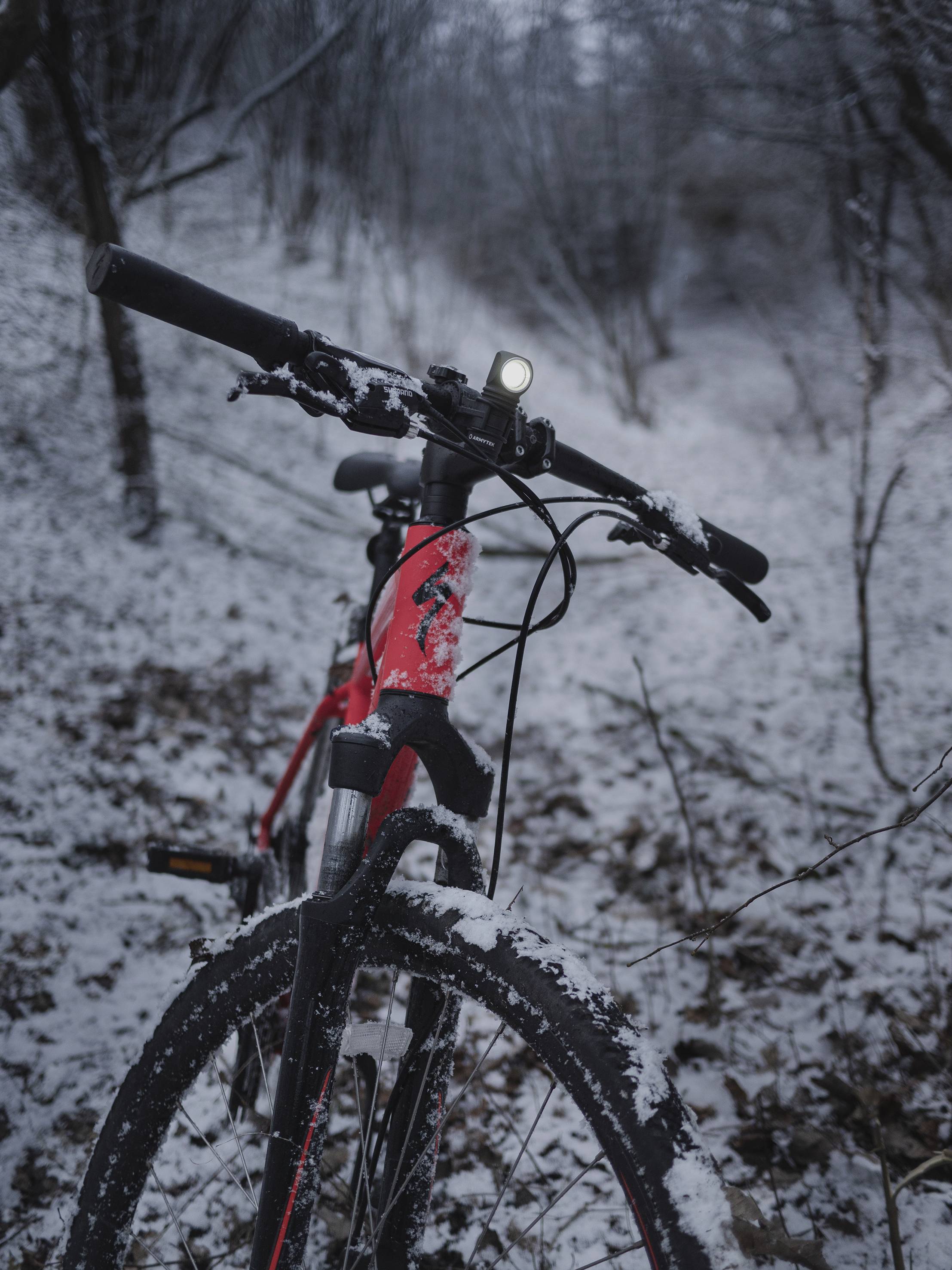 Ein rotes Mountainbike steht auf einem verschneiten Waldweg. Es ist Winter, und das Fahrradlicht leuchtet.
