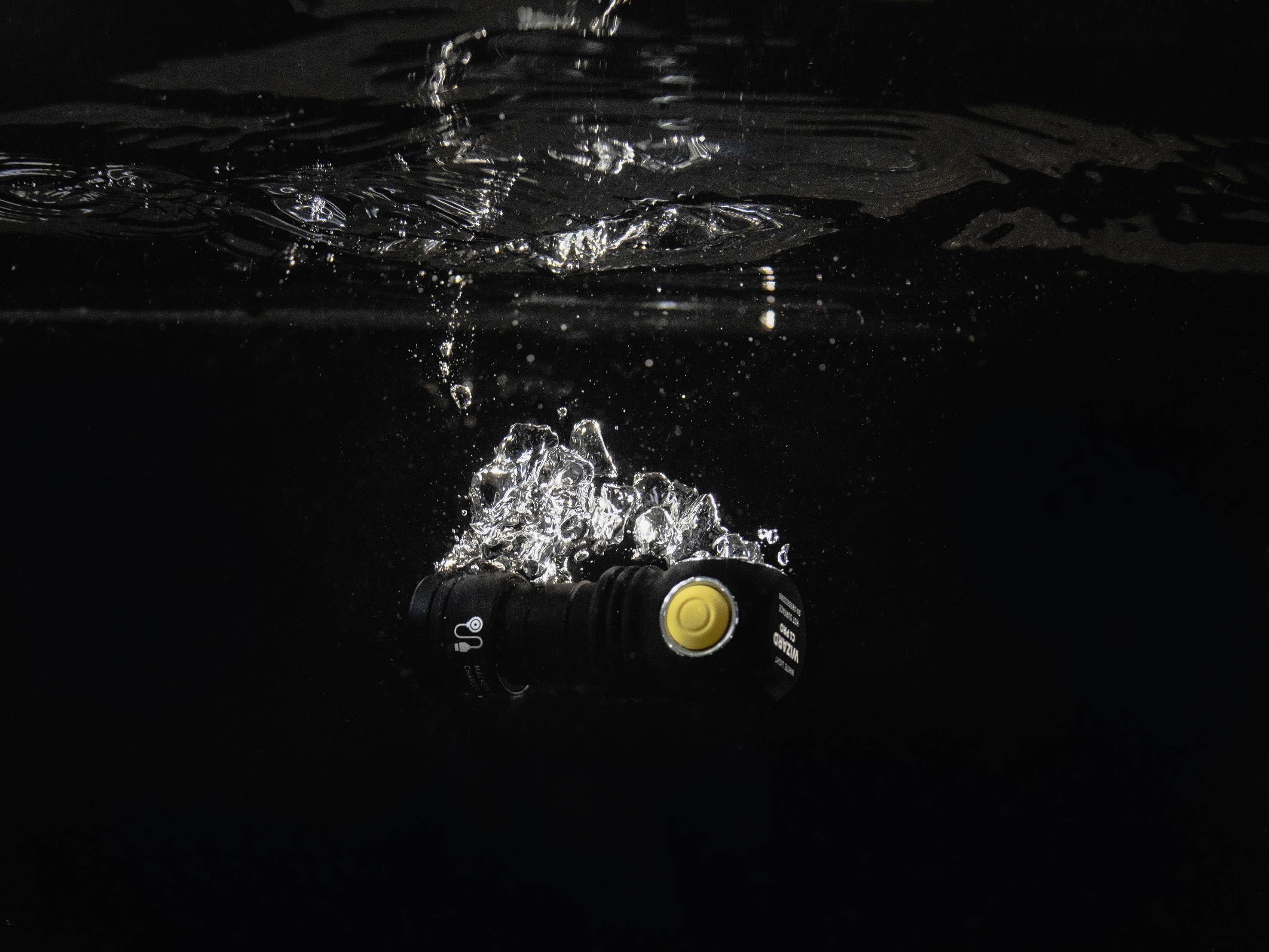Eine schwarze Taschenlampe mit einem gelben Knopf sinkt durch dunkles Wasser, umgeben von sprudelnden Luftblasen.