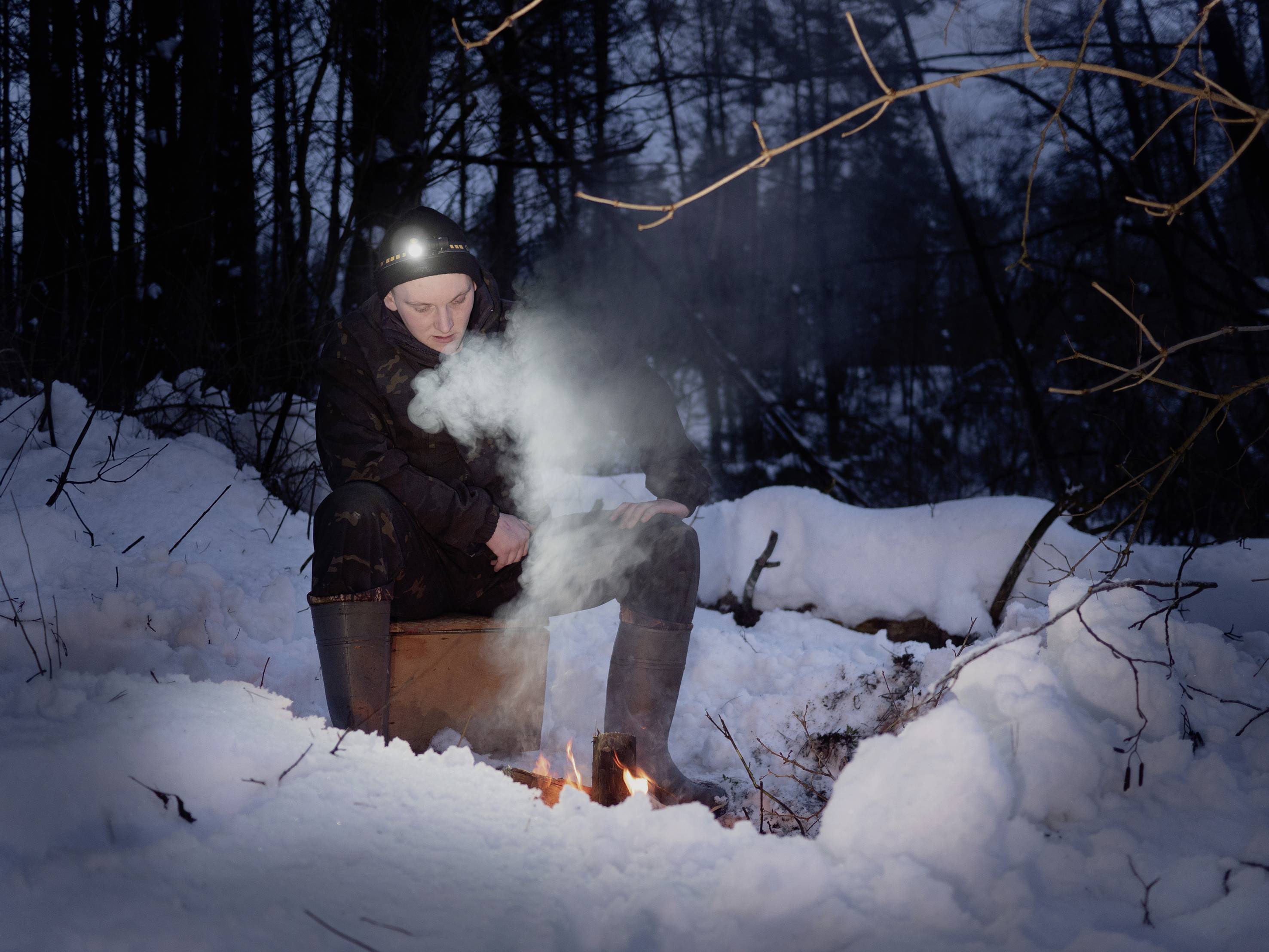 Eine Person sitzt im Schneewald bei Nacht an einem kleinen Feuer und trägt eine Stirnlampe. Es ist ruhig und atmosphärisch.