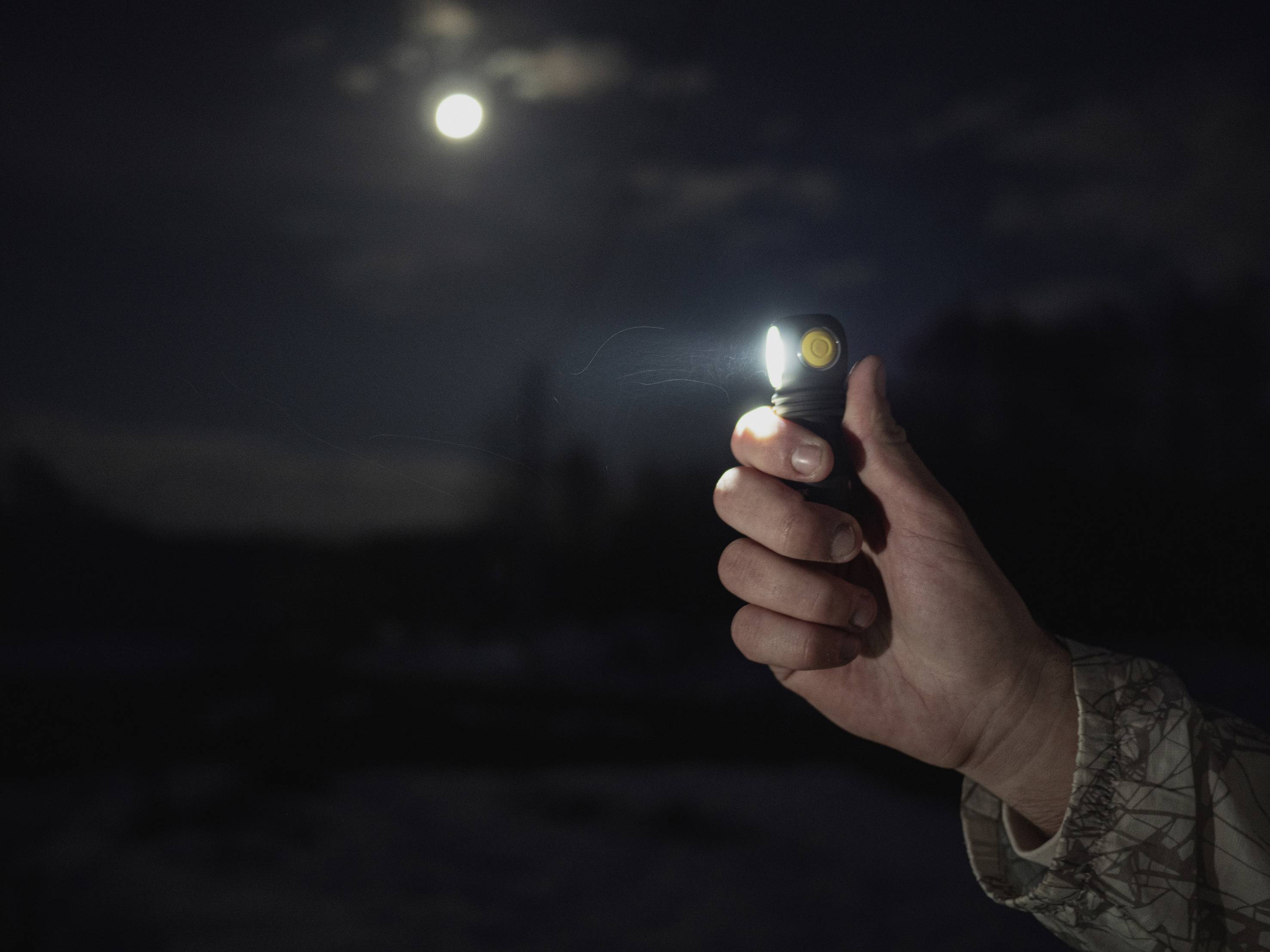Eine Hand hält eine leuchtende Taschenlampe in der Dunkelheit, während der Vollmond im Hintergrund am Himmel steht.