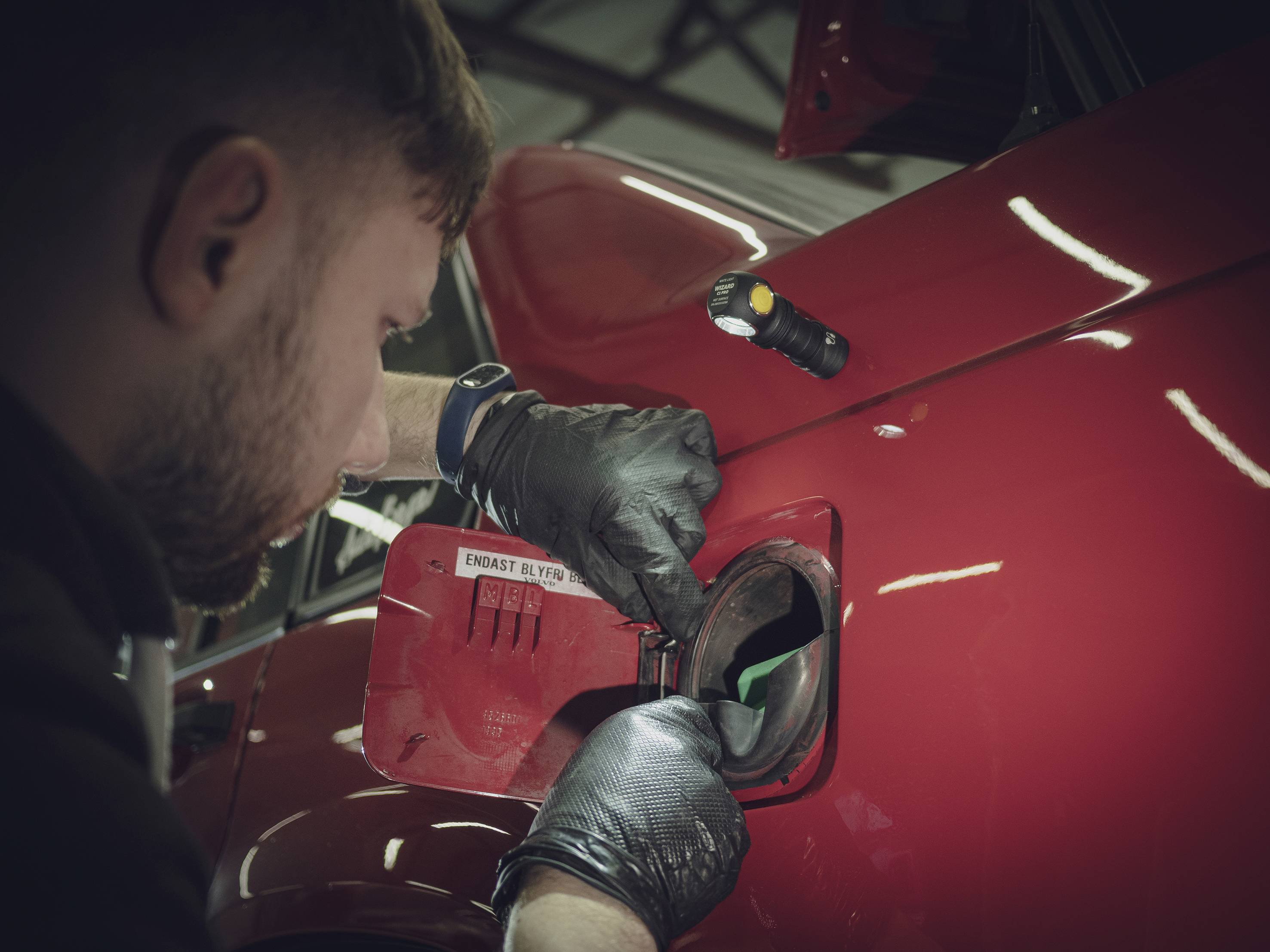 Ein Mann tankt ein rotes Auto. Er trägt schwarze Handschuhe und verwendet eine Taschenlampe, um die Tanköffnung zu beleuchten.