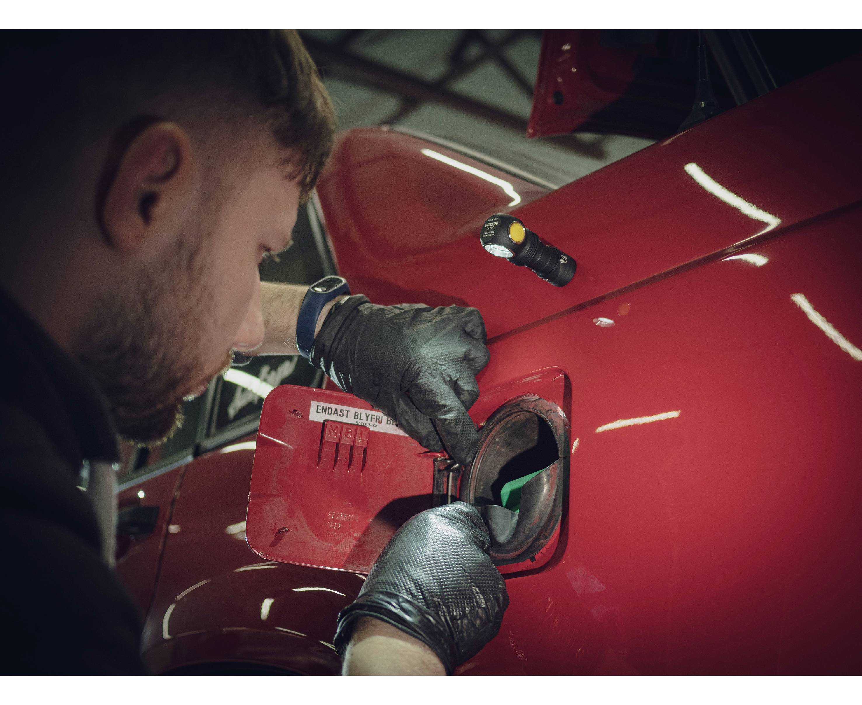 Ein Mechaniker mit schwarzen Handschuhen überprüft den geöffneten Tankdeckel eines roten Autos in einer Werkstatt.