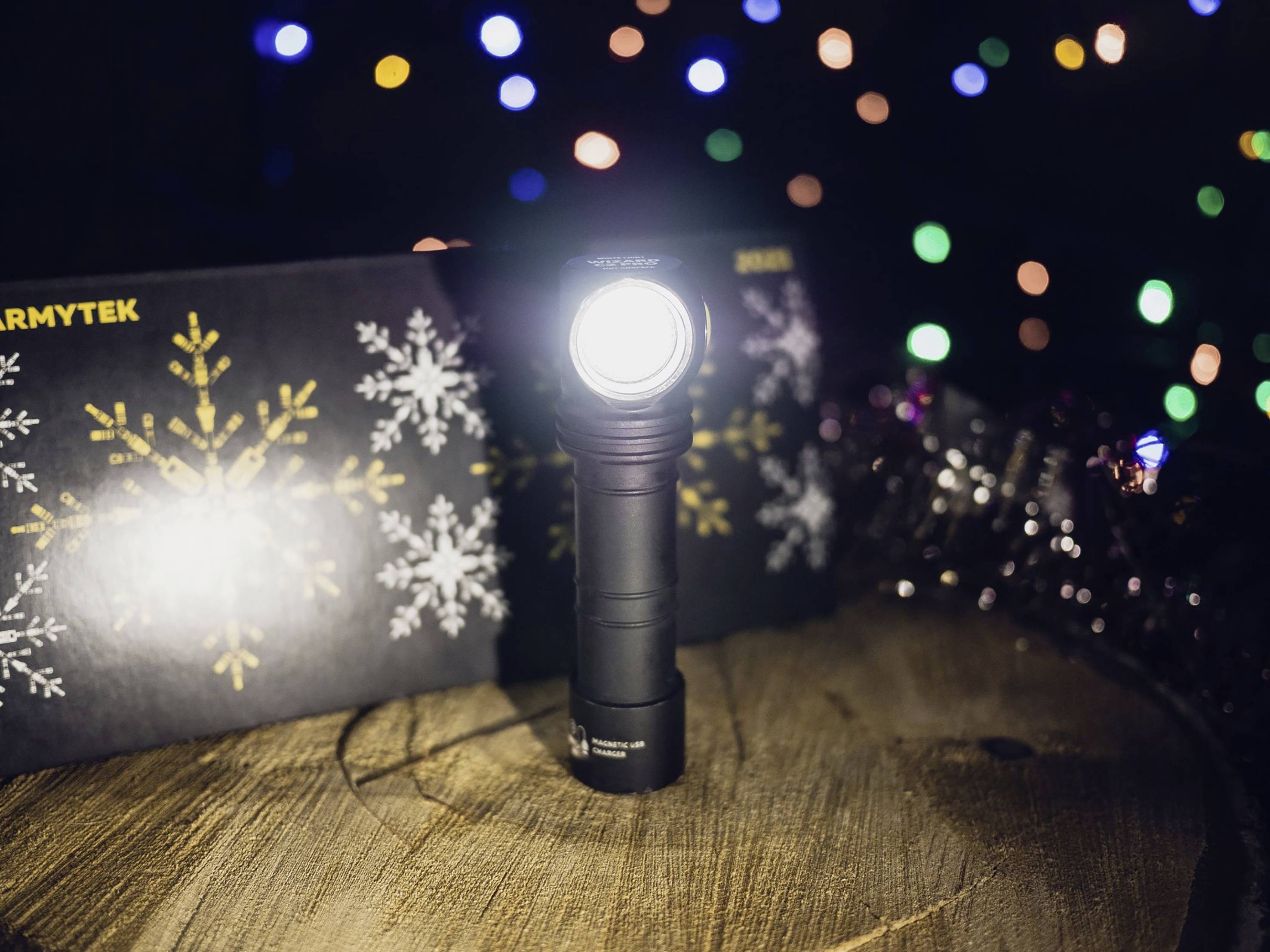 Eine schwarze Taschenlampe leuchtet auf einem Holztisch. Im Hintergrund sind bunte, unscharfe Lichter und ein schwarzer Karton mit Schneeflocken.