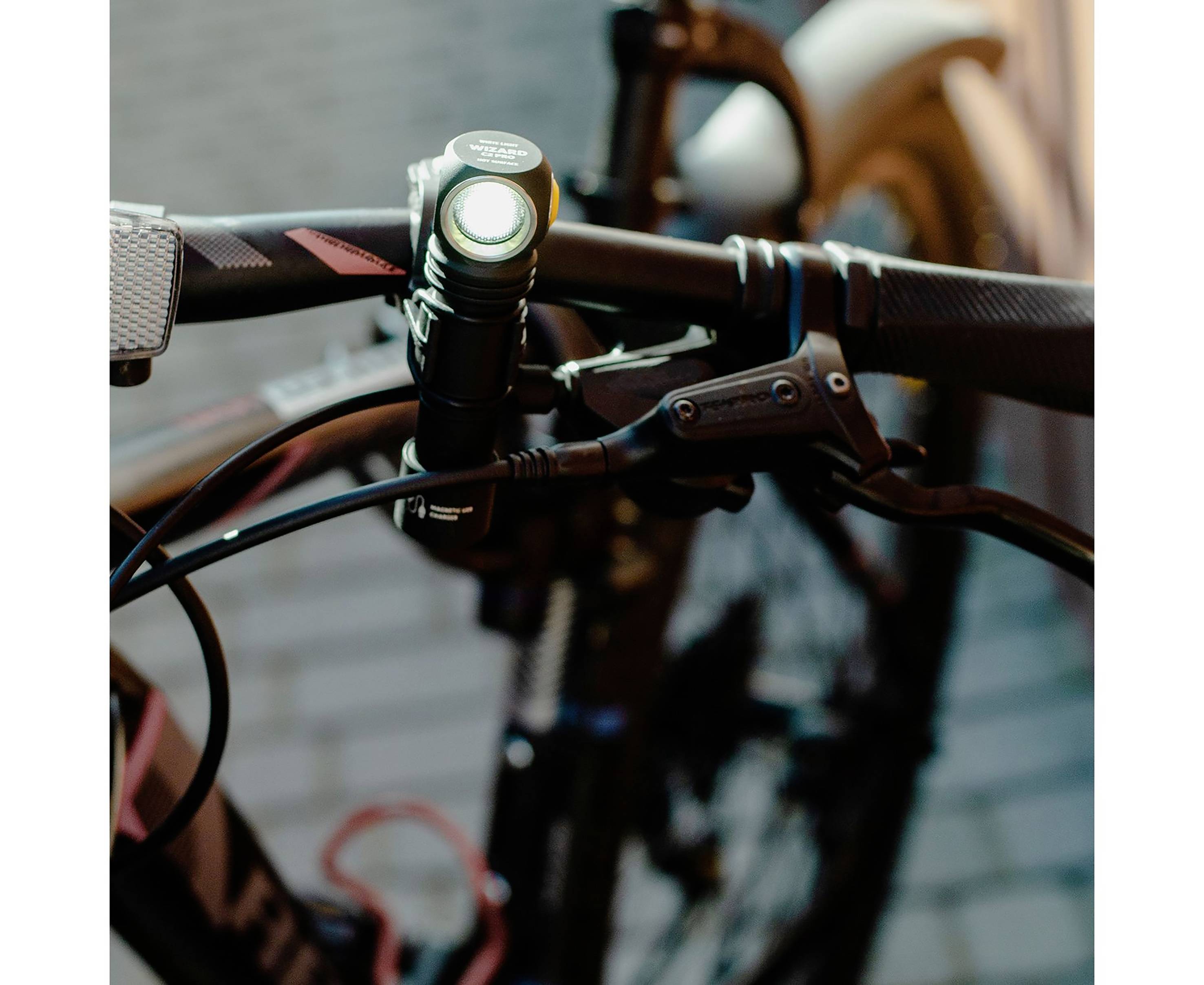 Nahaufnahme eines Fahrradlenkers mit einer montierten Taschenlampe, die eingeschaltet ist. Im Hintergrund verschwommene Pflastersteine.