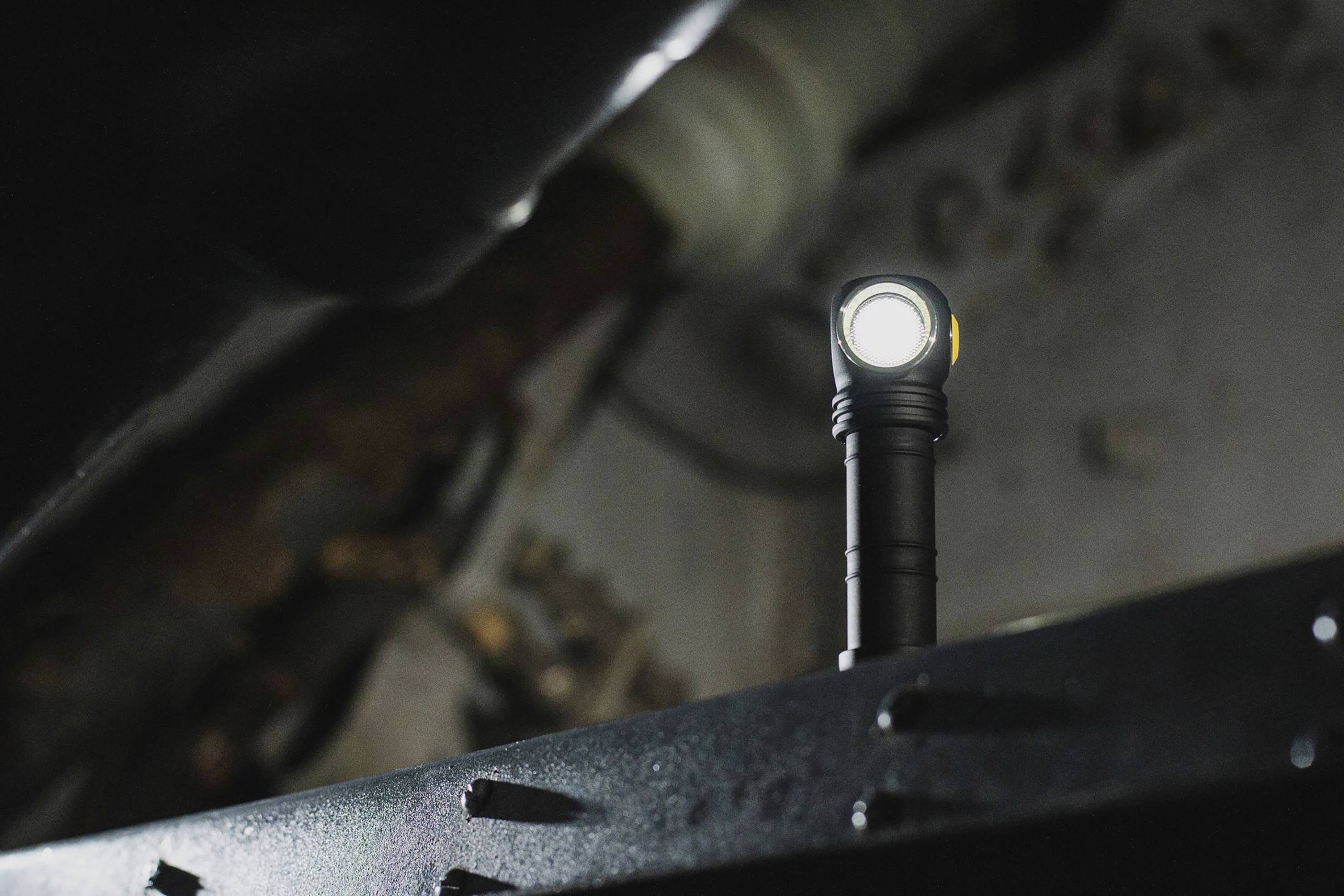 Taschenlampe mit hellem Licht, steht auf einer Metalloberfläche in einem dunklen Raum. Hintergrund unscharf, Details des Raums kaum erkennbar.