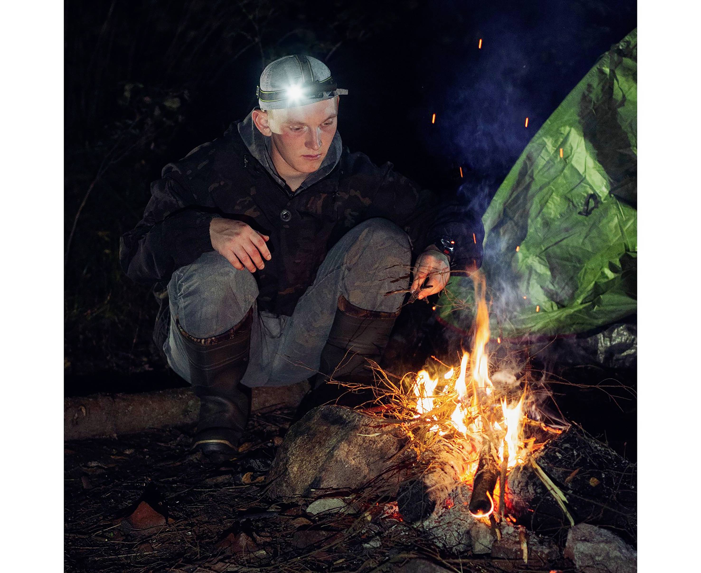 Eine Person hockt im Dunkeln neben einem Lagerfeuer und trägt eine Stirnlampe. Sie ist in einem Wald und es gibt einen grünen Hintergrund.