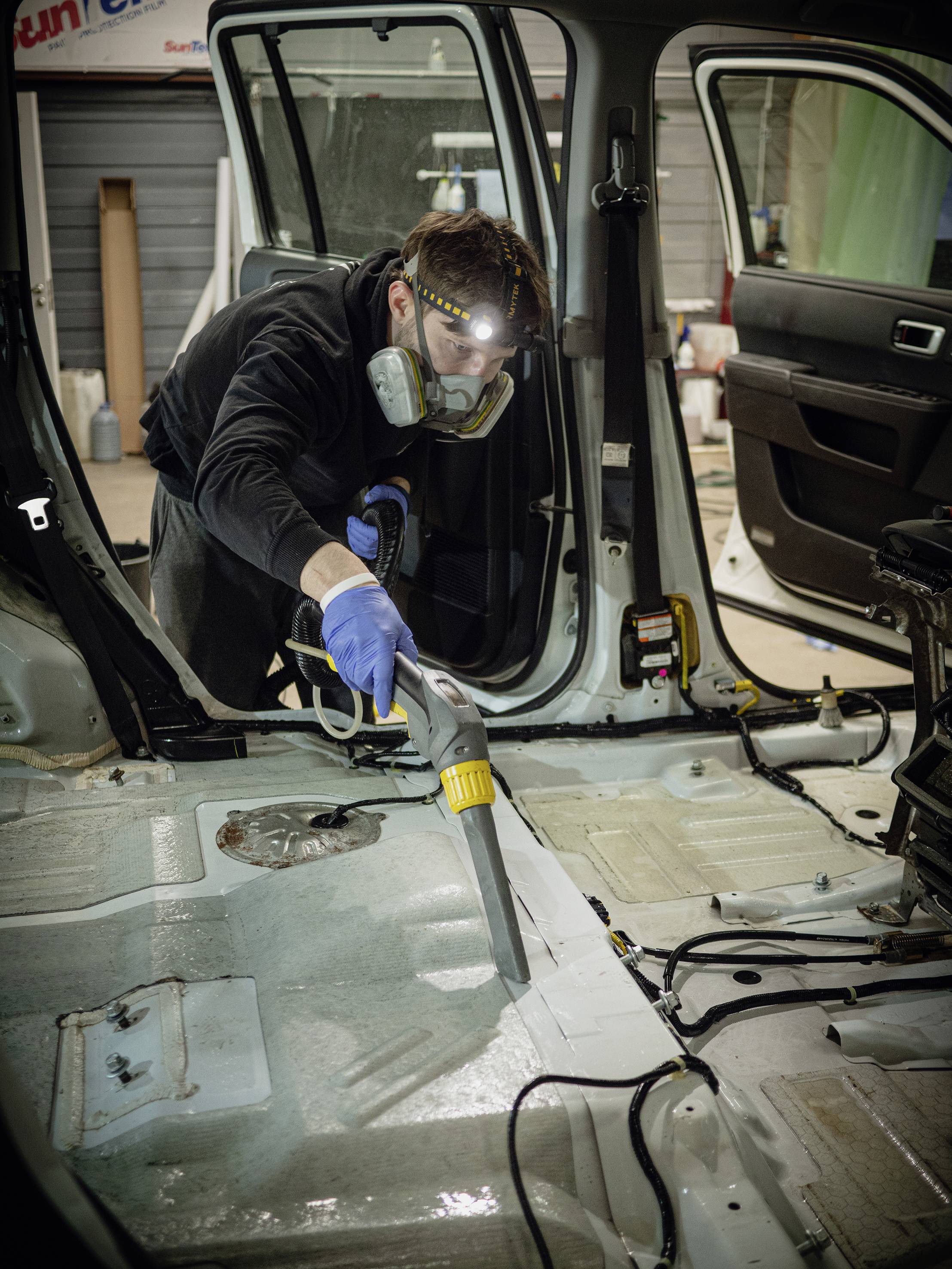 Eine Person, die mit Schutzmaske und -handschuhen einen Staubsauger im Innenraum eines fahrzeuglosen Autos verwendet.