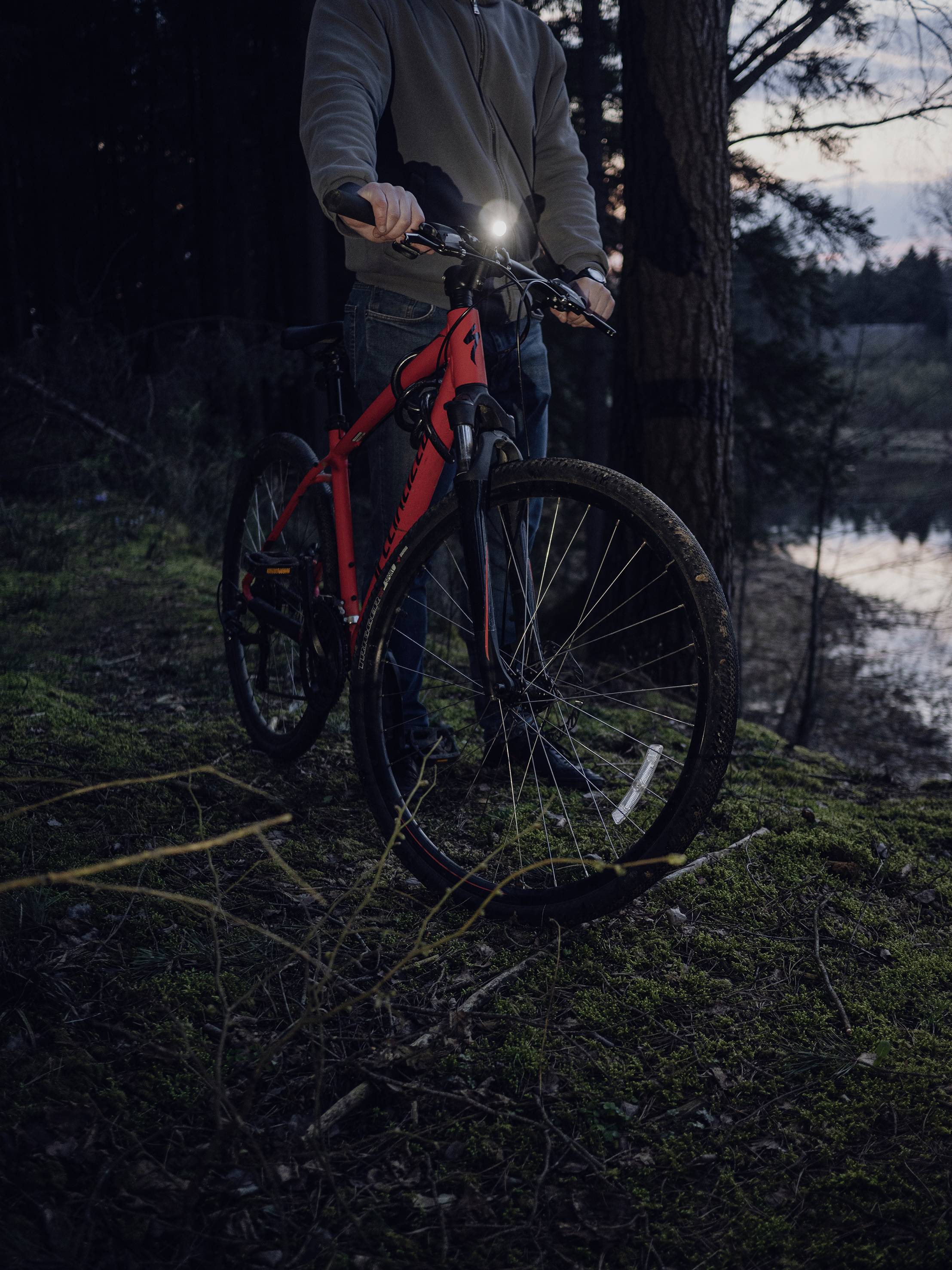Eine Person steht im Wald mit einem roten Fahrrad. Das Licht des Fahrrads ist an, es wird Abend, und der Himmel ist leicht beleuchtet.