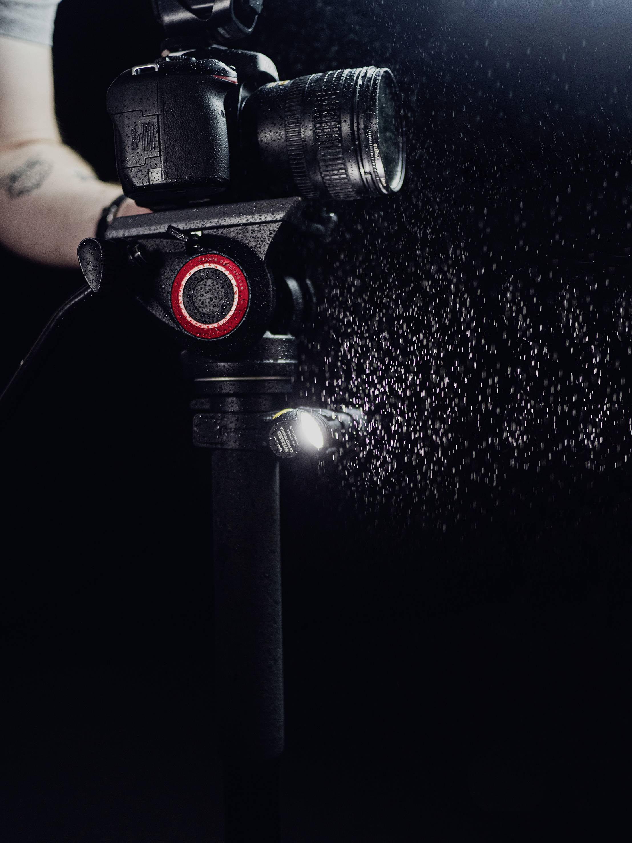 Kamera auf einem Stativ, von schräg oben fotografiert. Eine Hand justiert die Einstellungen, während Wassertropfen im Hintergrund sichtbar sind.