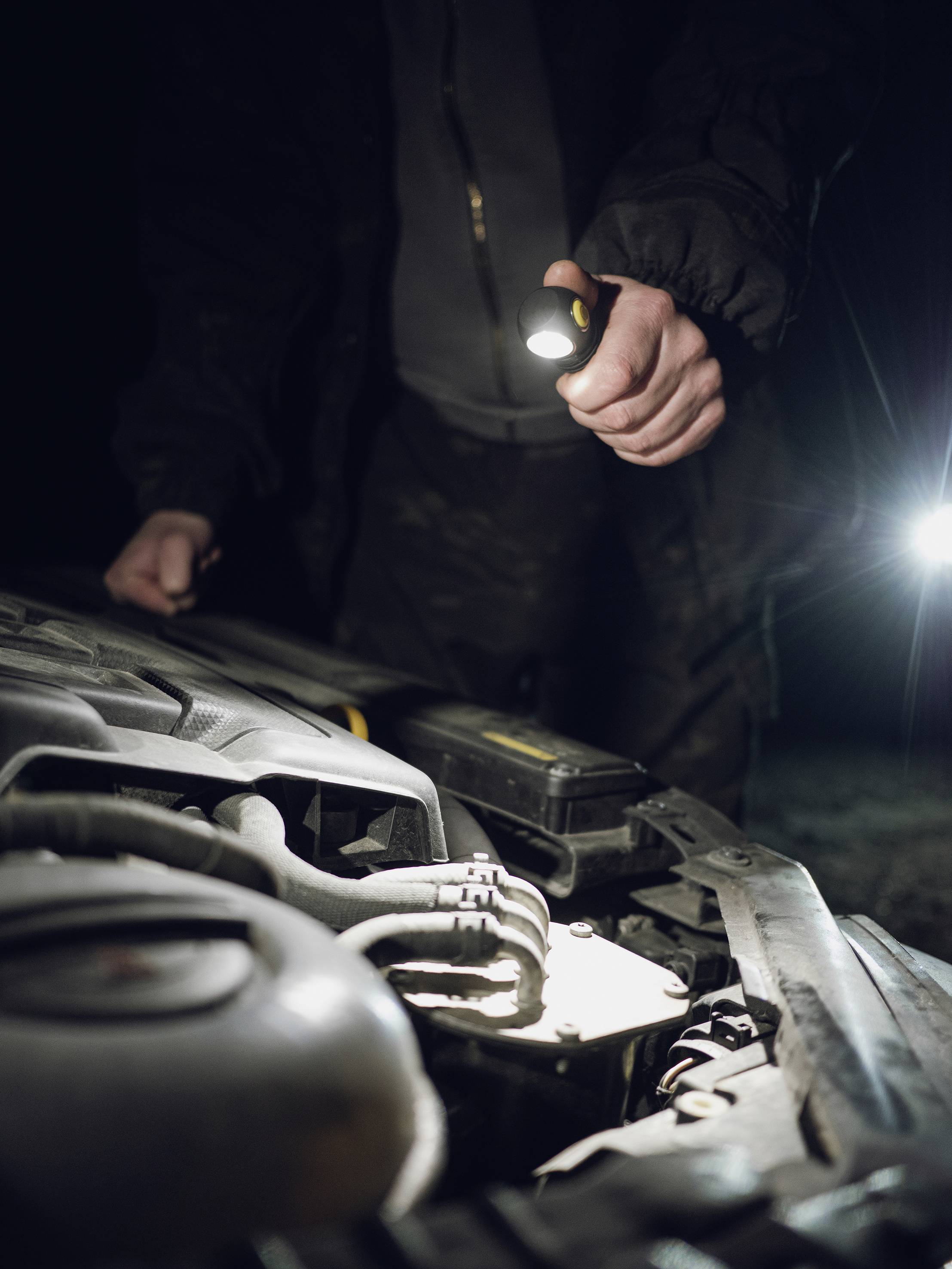 Ein Mensch untersucht den Motorraum eines Autos bei Nacht mit einer Taschenlampe.