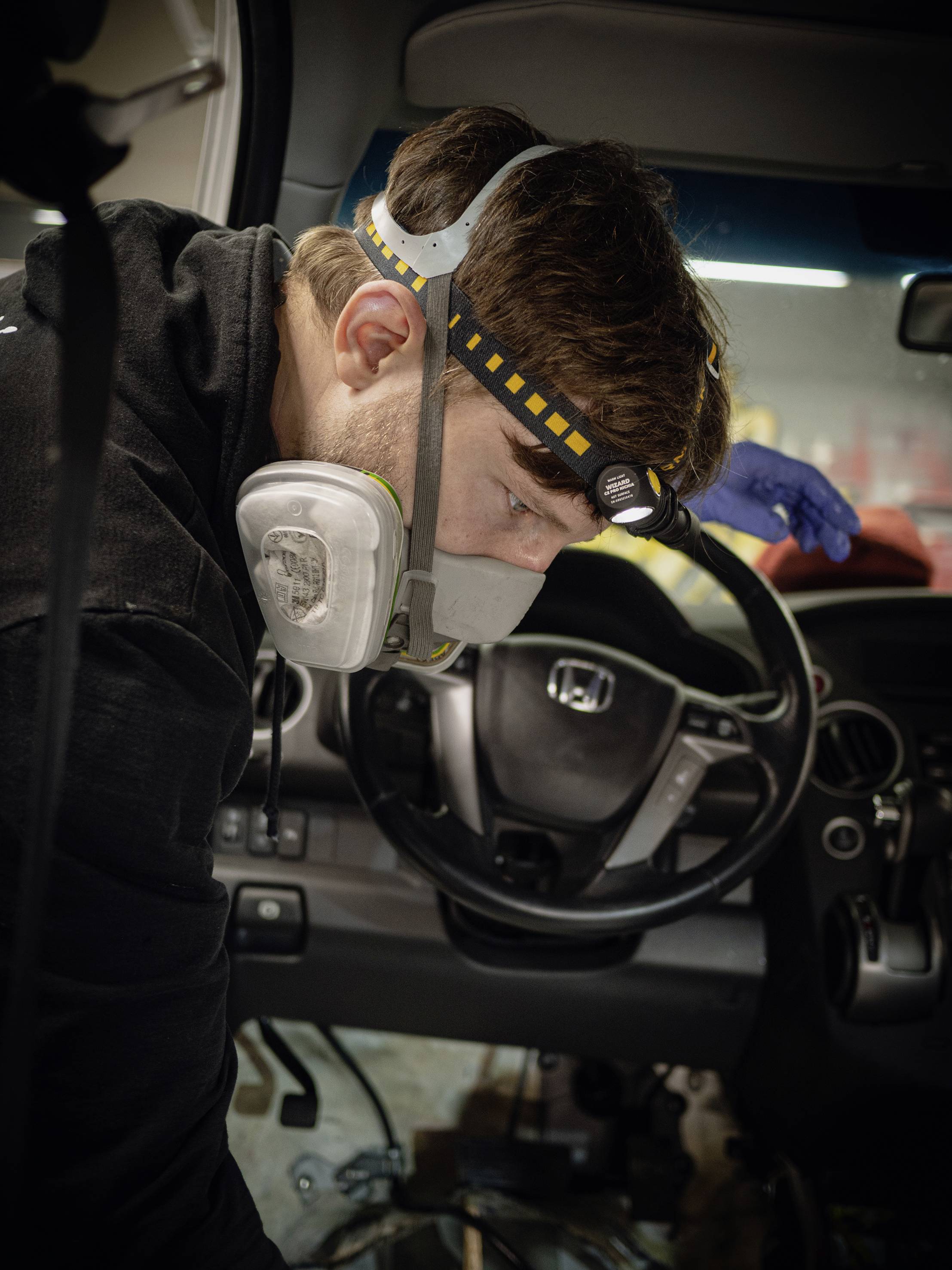 Ein Mann trägt Atemschutzmaske und Stirnlampe, arbeitet konzentriert im Inneren eines Autos, vermutlich an einem technischen Problem.