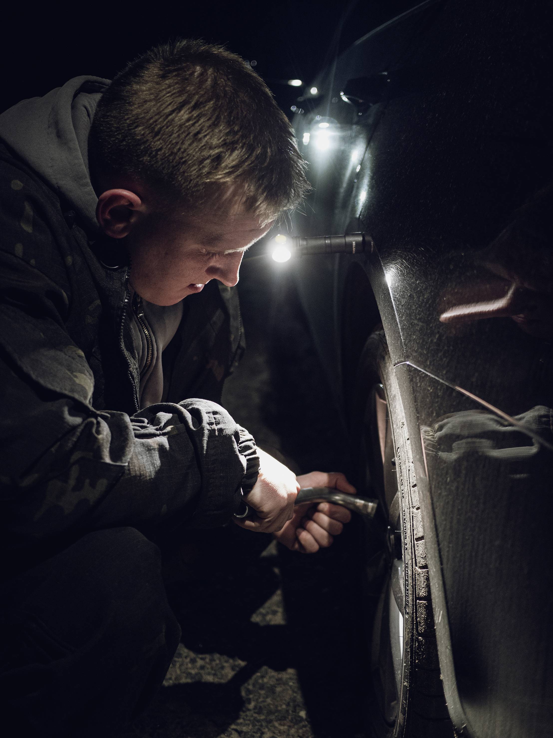 Ein Mann wechselt nachts auf einer dunklen Straße den Reifen eines Autos. Eine Taschenlampe beleuchtet die Arbeit.