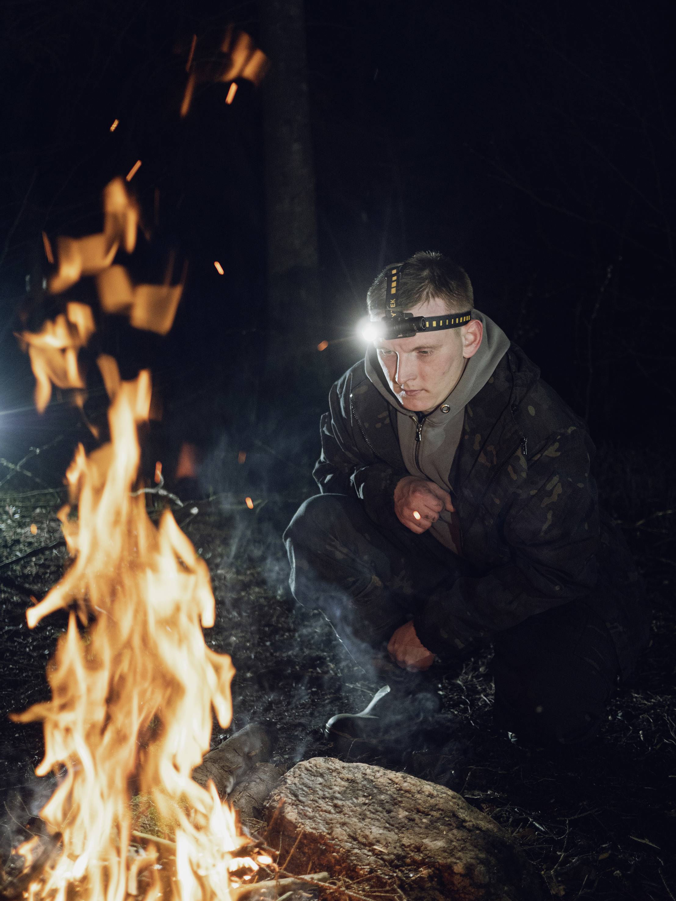 Eine Person mit Stirnlampe hockt nachts am Lagerfeuer im Wald, umgeben von Bäumen.
