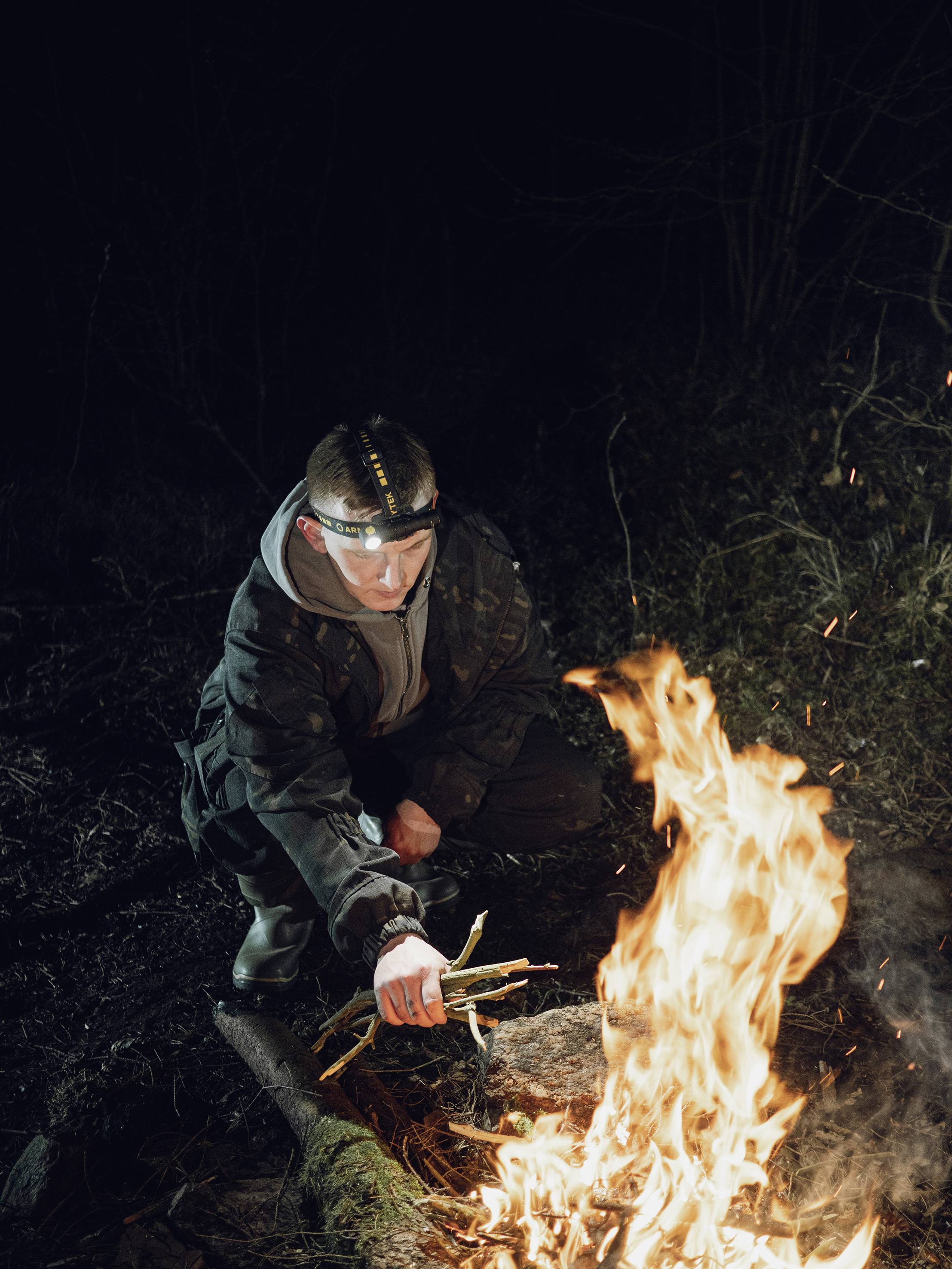 Eine Person in Outdoor-Kleidung zündet bei Nacht mit einem Feuerstarter ein Lagerfeuer an.
