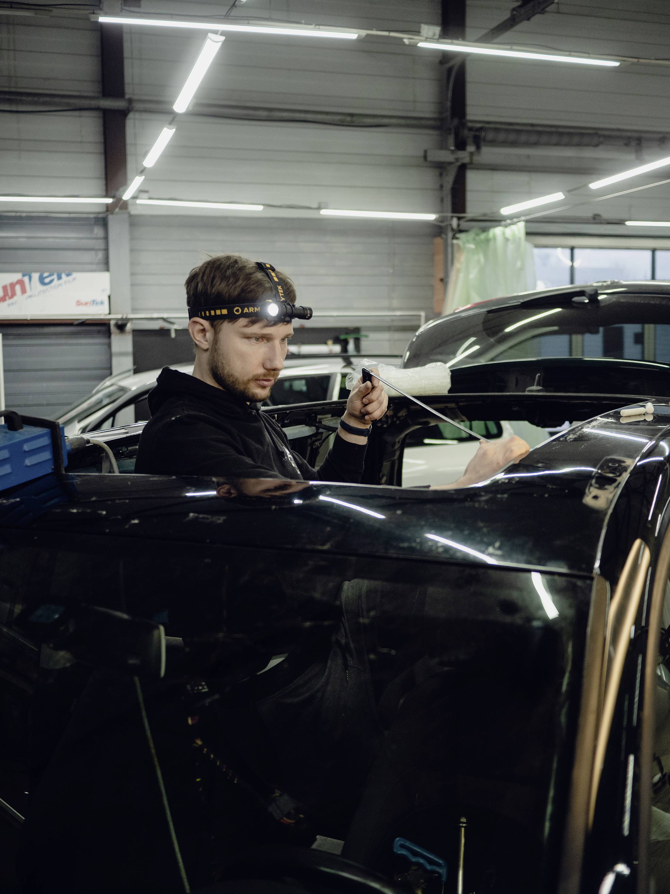 Ein Mann arbeitet in einer Autowerkstatt. Er trägt ein Stirnlicht und repariert ein schwarzes Auto in einer Werkstattumgebung.
