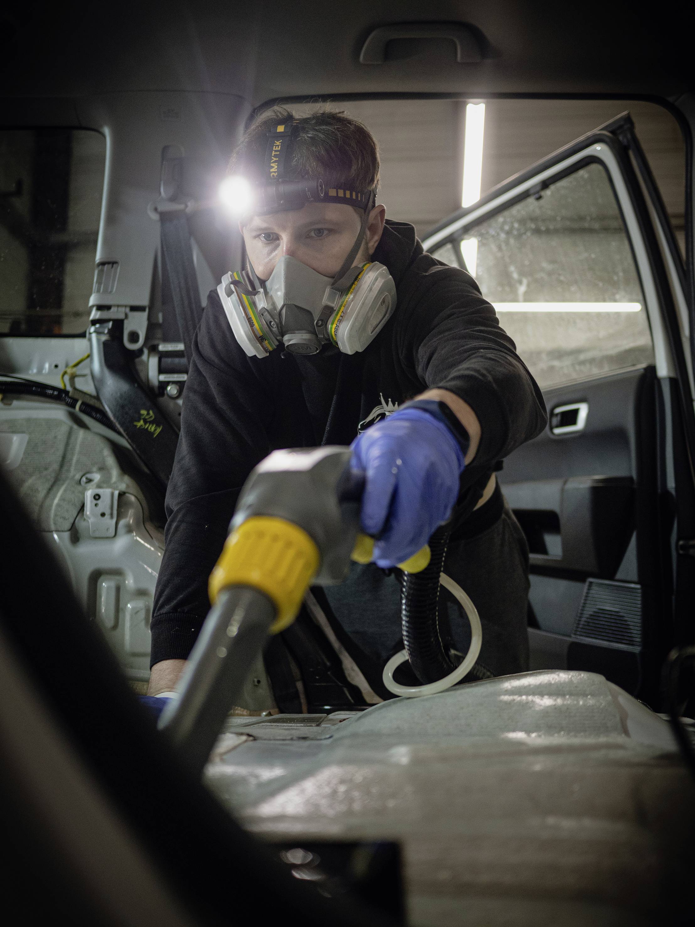 Ein Mann reinigt den Innenraum eines Autos mit einer Maschine. Er trägt einen Atemschutz, Handschuhe und eine Stirnlampe, um die Arbeit zu erleichtern.