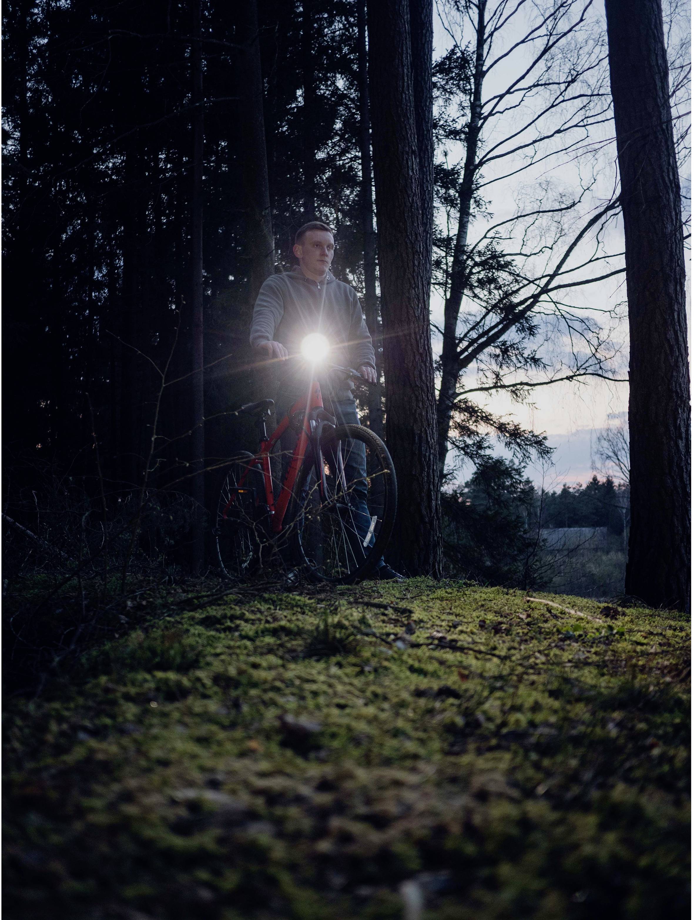 Eine Person steht mit einem Fahrrad im Wald, das Vorderlicht des Fahrrads ist eingeschaltet. Es ist Abenddämmerung.