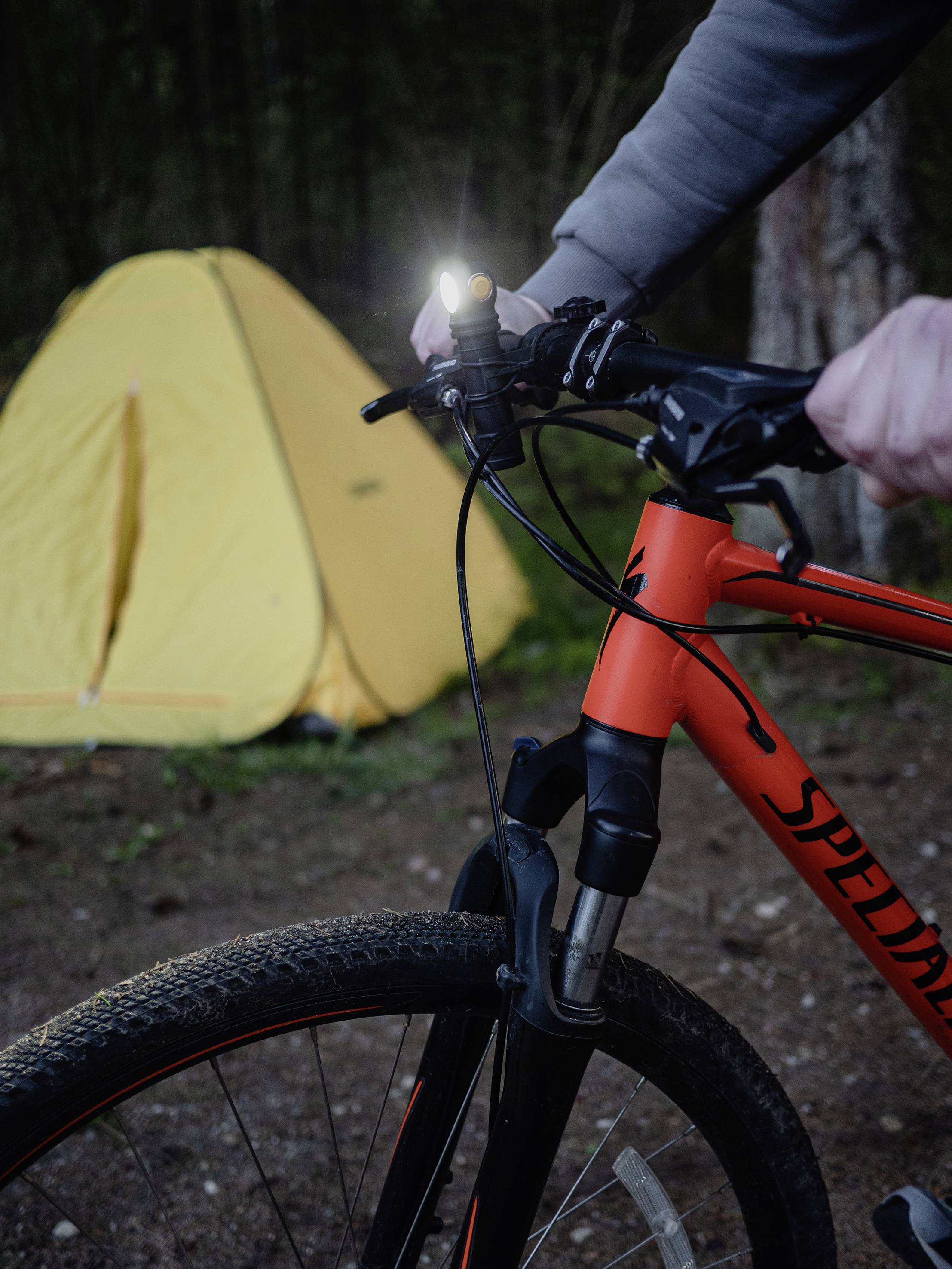 Eine Person hält ein Fahrradlenker mit eingeschaltetem Licht. Im Hintergrund steht ein gelbes Zelt. Die Szene spielt im Freien.