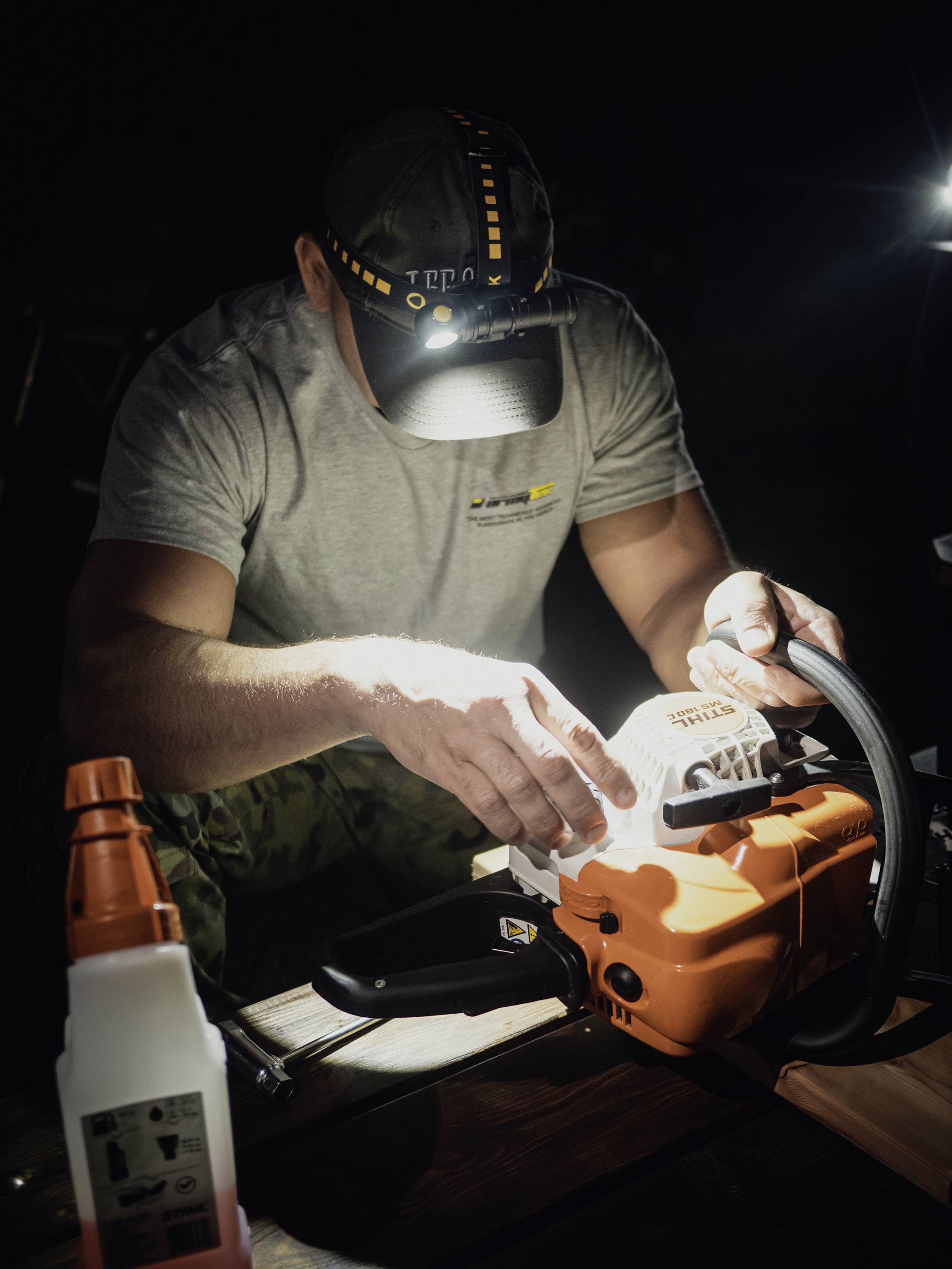 Ein Mann mit Stirnlampe arbeitet nachts an einer Motorsäge, die auf einem Tisch liegt, und benutzt eine Feile zur Wartung der Kette.