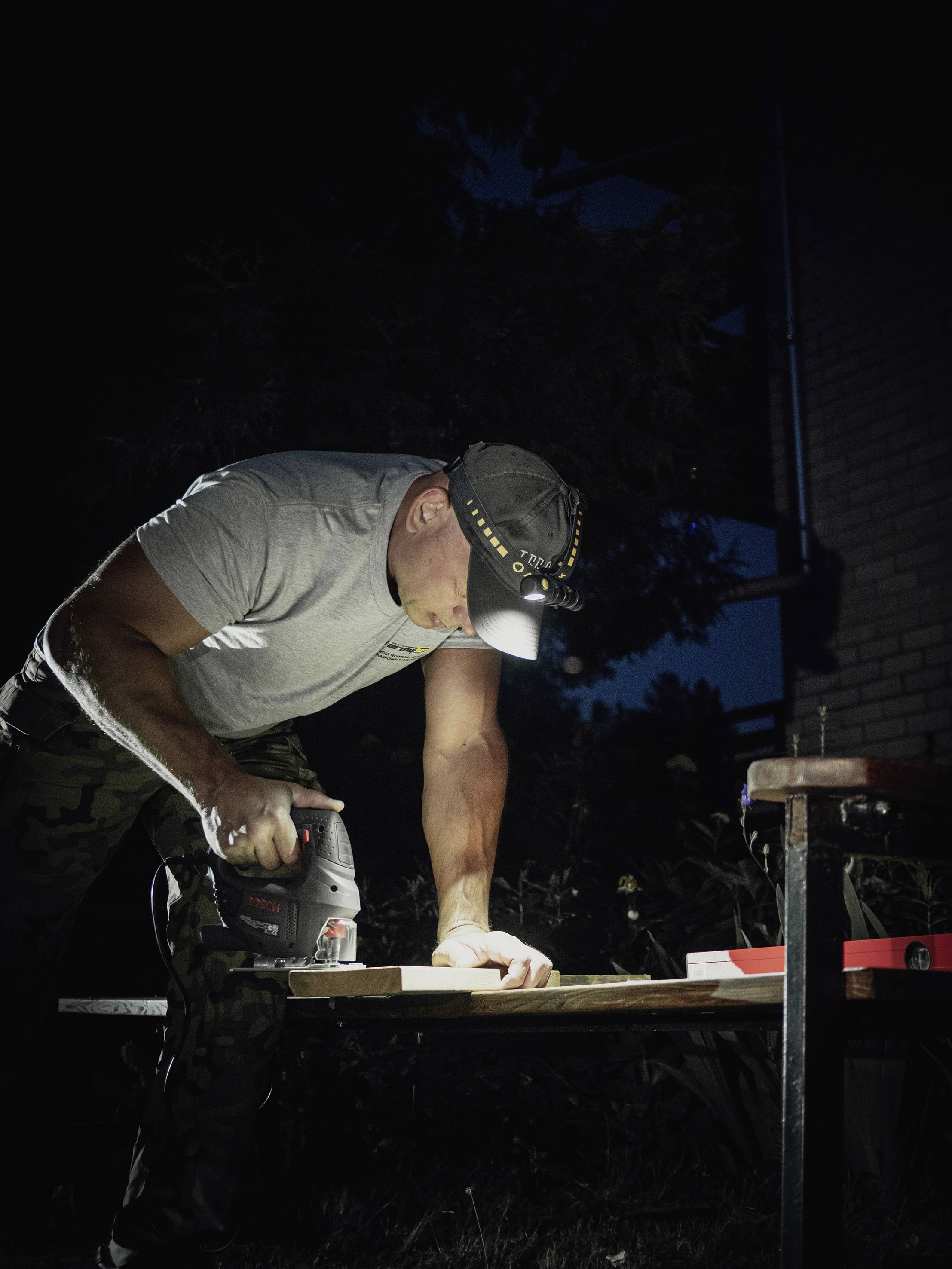 Ein Mann mit einer Stirnlampe sägt nachts Holz auf einem Tisch im Freien. Seine Konzentration liegt auf der Arbeit.