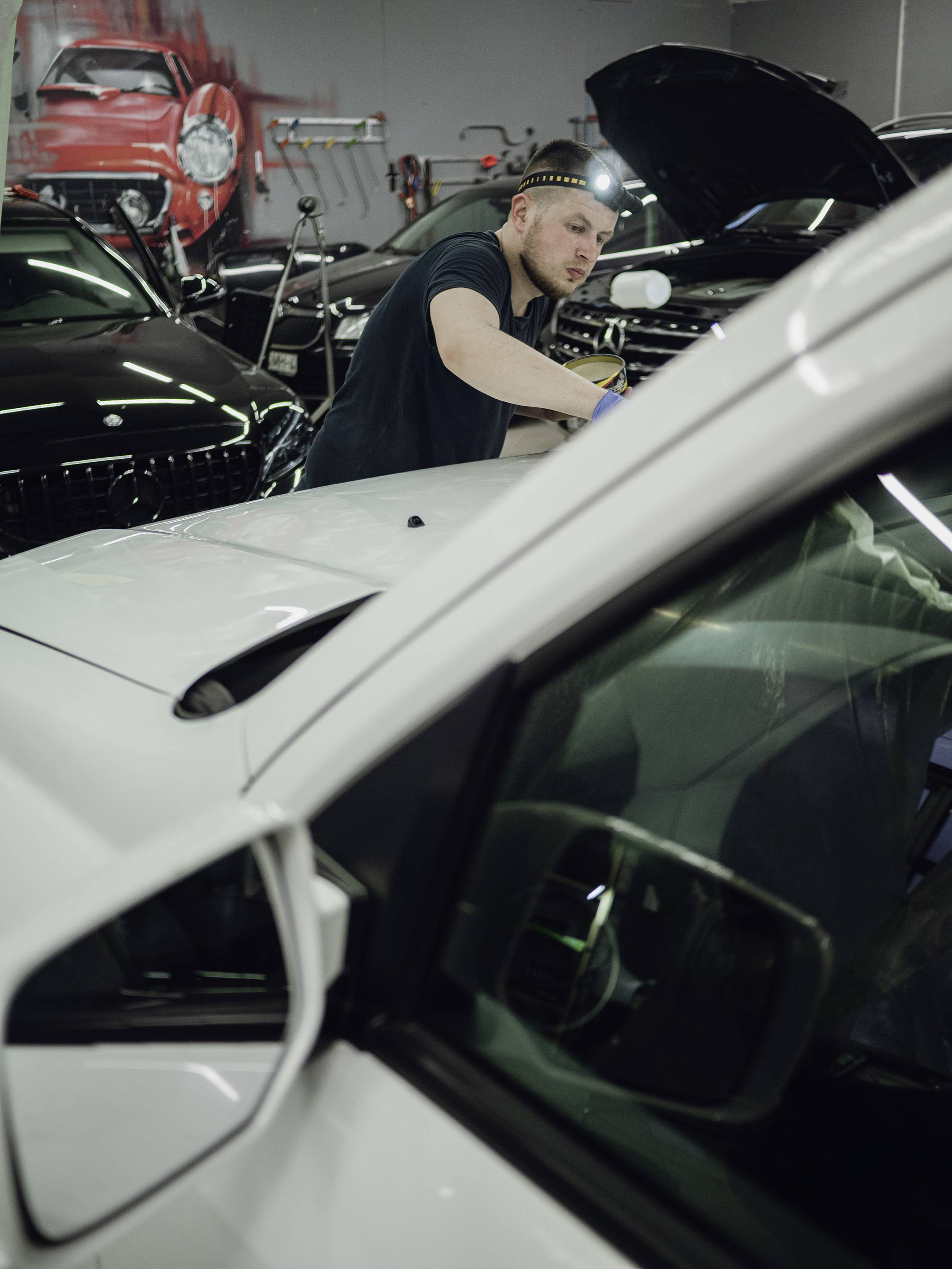 Mann repariert ein Auto in einer Werkstatt. Er trägt ein Stirnbandlicht und arbeitet konzentriert an der Vorderseite des Fahrzeugs.